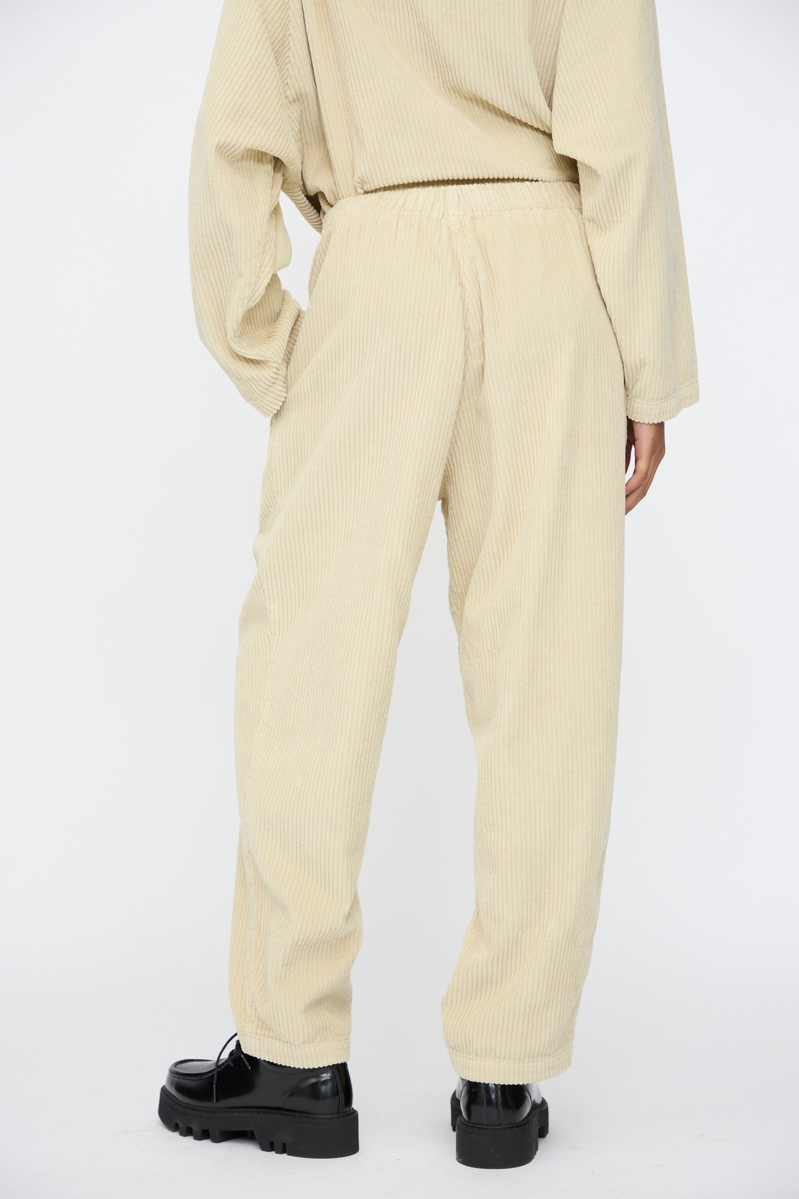 A person wears Black Crane Carpenter Pants in Ivory—relaxed fit cotton corduroy with an elastic waist—paired with a matching top and black chunky shoes, standing with their back to the camera.