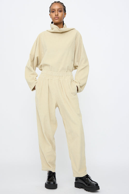 A person stands against a white background wearing Black Crane Carpenter Pants in Ivory—relaxed fit cotton corduroy with an elastic waist—paired with a matching high-collar top, black shoes, hands in pockets, and a neutral expression.