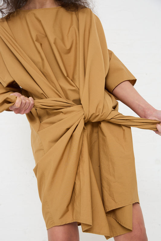 A person is adjusting an Organic Cotton Bow Dress in Camel by Black Crane with a knotted detail at the waist.