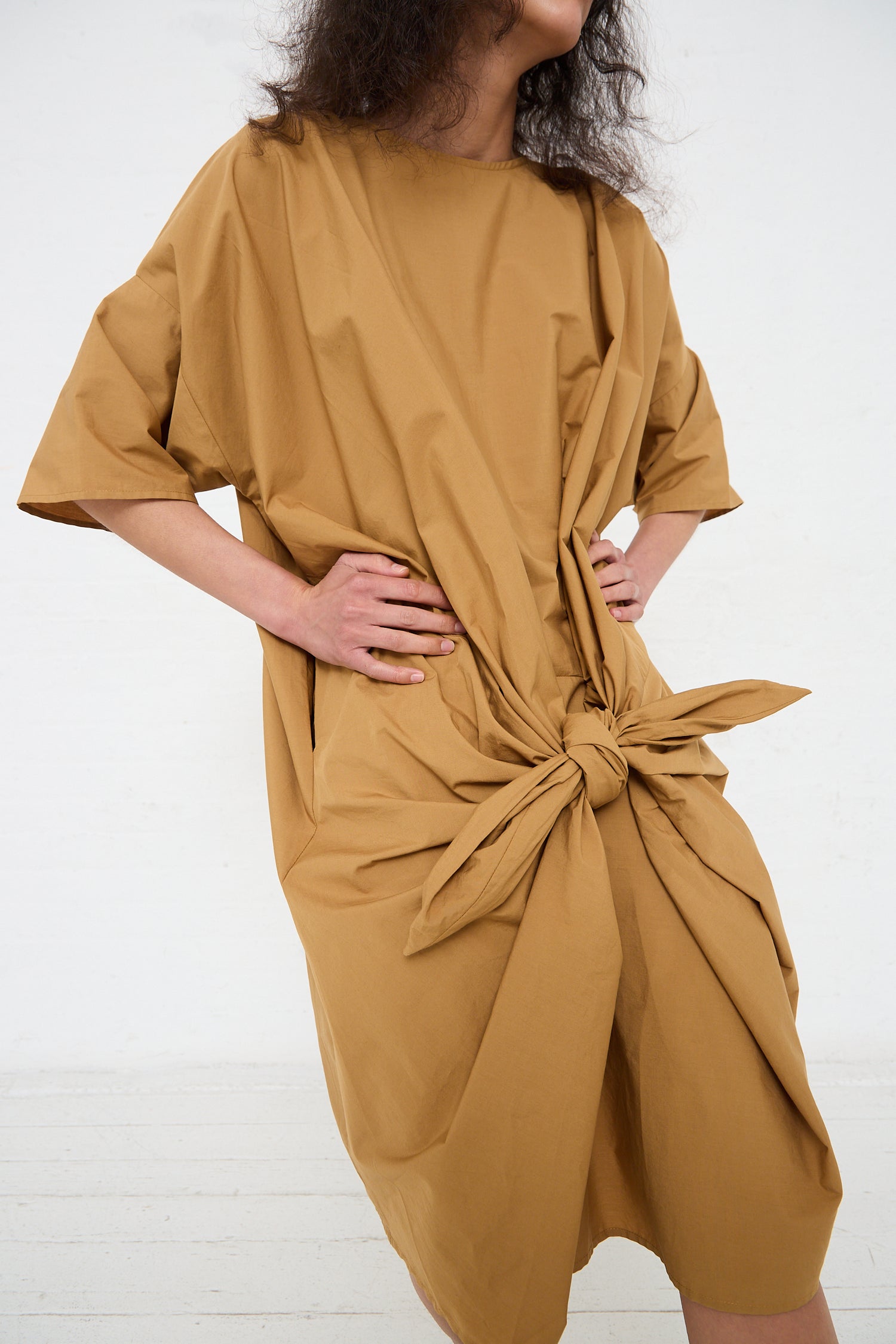Woman in a Black Crane Organic Cotton Bow Dress in Camel standing against a white background.