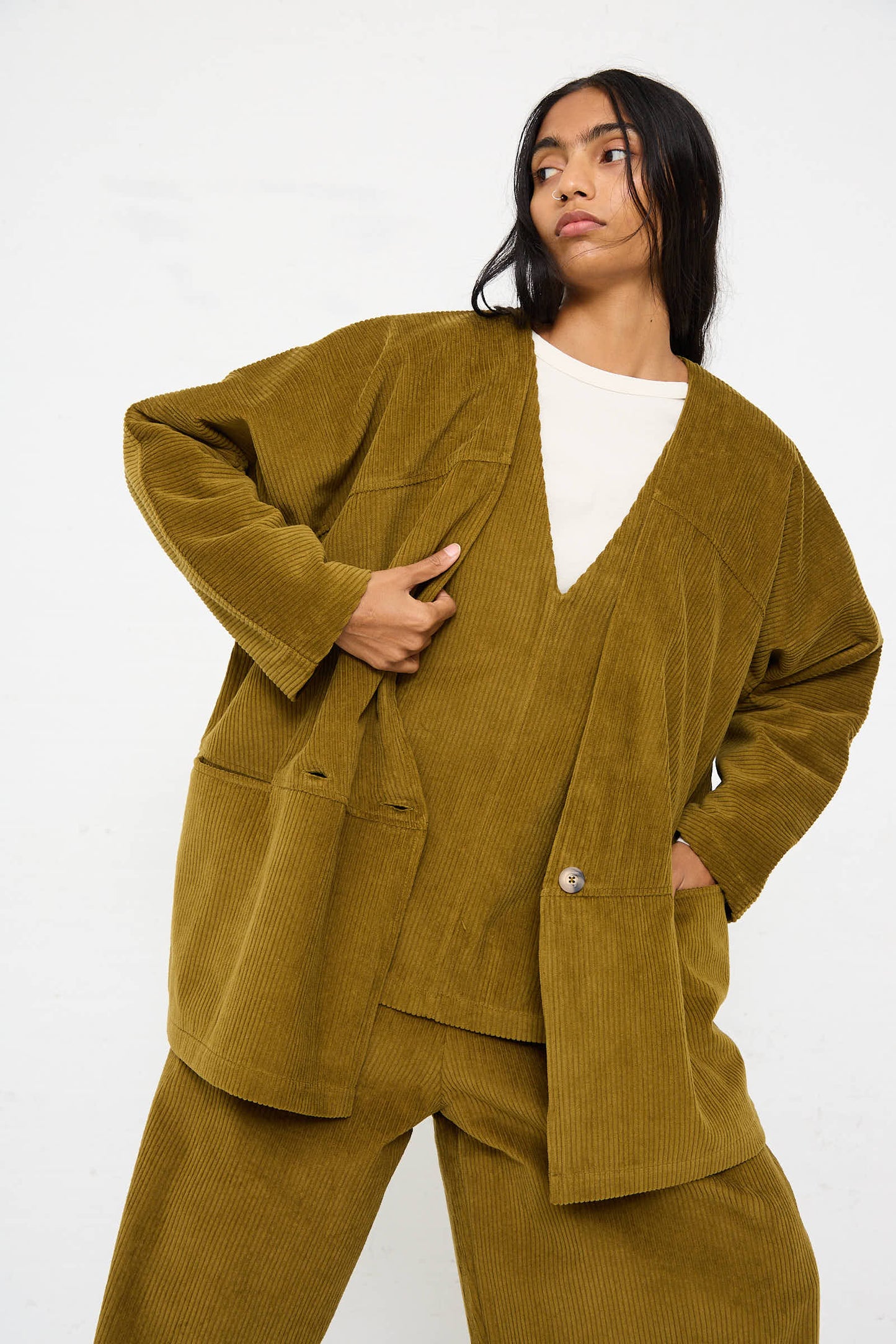 A person wearing a dark olive, heavy cotton corduroy outfit from Black Crane, consisting of the Cotton Corduroy Geometric Jacket and matching relaxed fit pants, poses in front of a plain background.