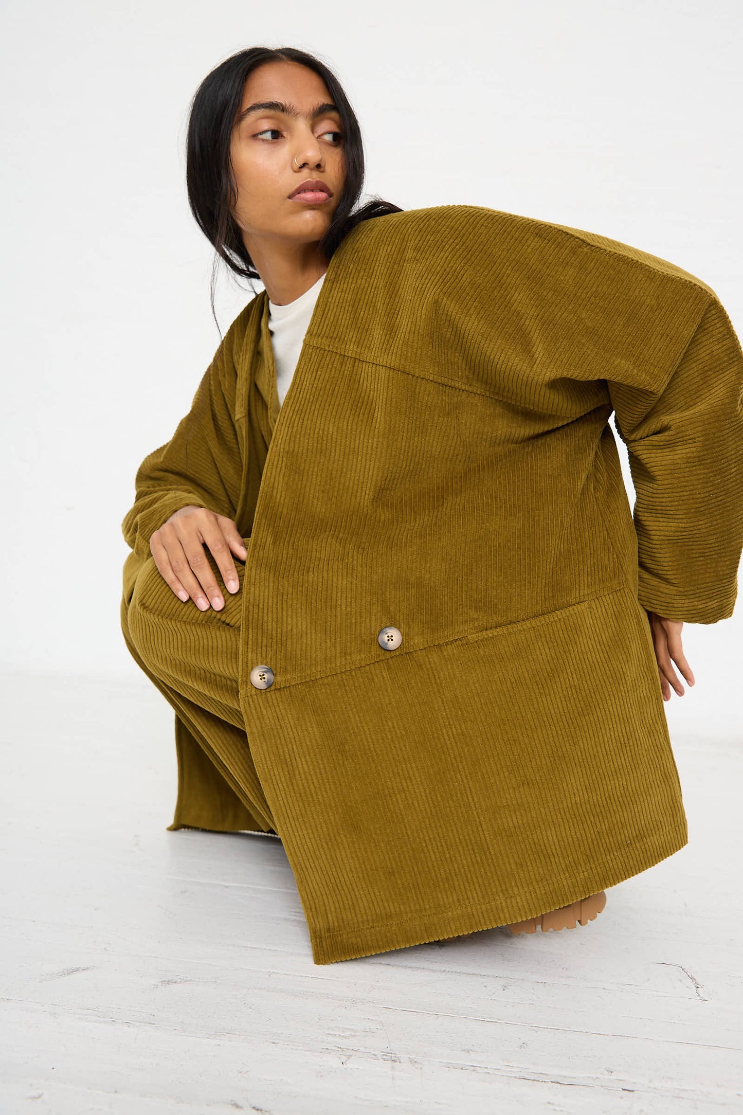 A person with long dark hair wearing the Black Crane Cotton Corduroy Geometric Jacket in Dark Olive, featuring an oversized and relaxed fit, is crouching on a white floor, looking to the side.