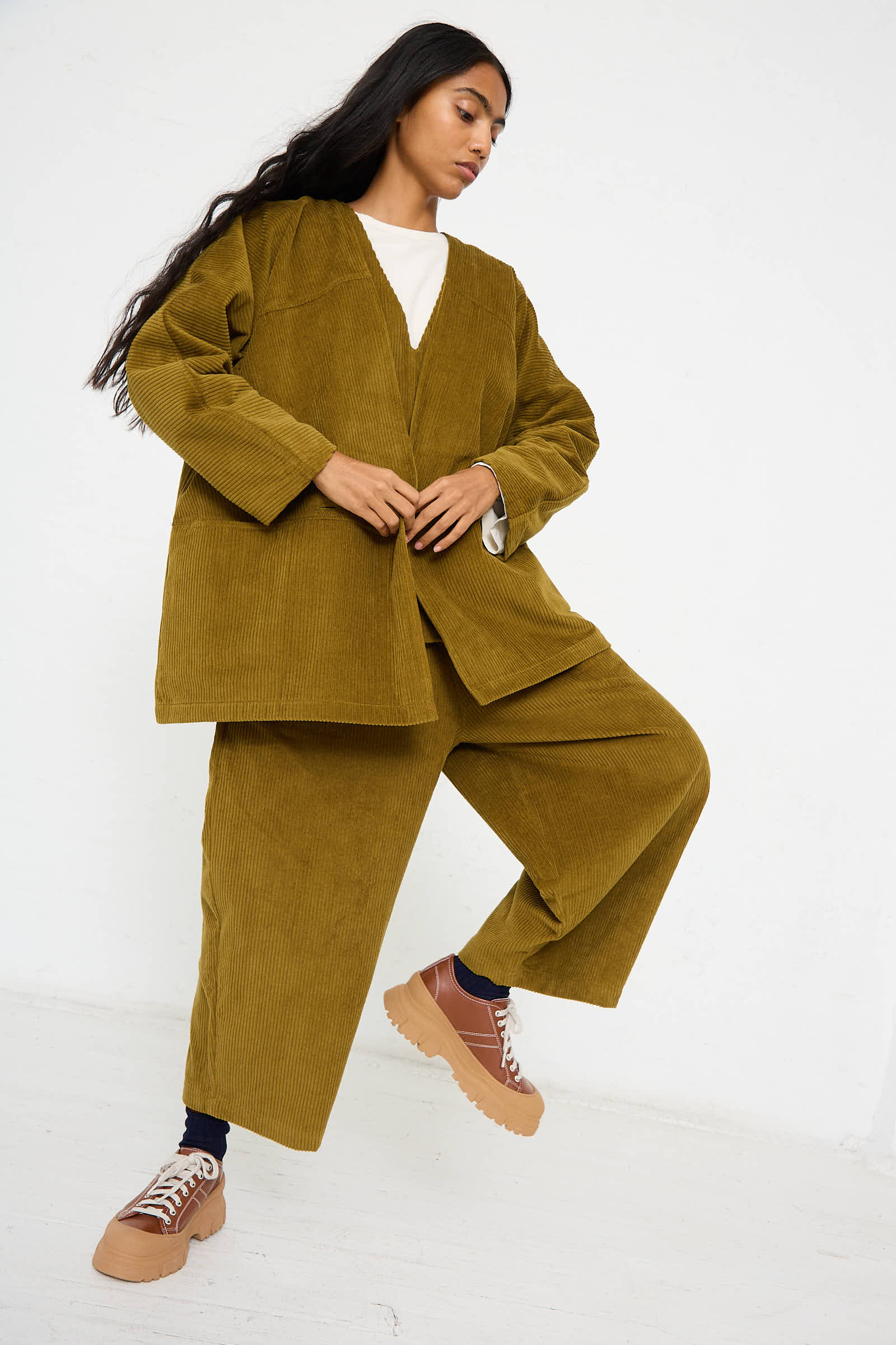 A person wearing an oversized Cotton Corduroy Geometric Jacket in Dark Olive by Black Crane, paired with a cream-colored inner shirt and brown platform shoes, poses against a plain white background. The relaxed fit gives an effortlessly chic vibe to the ensemble.