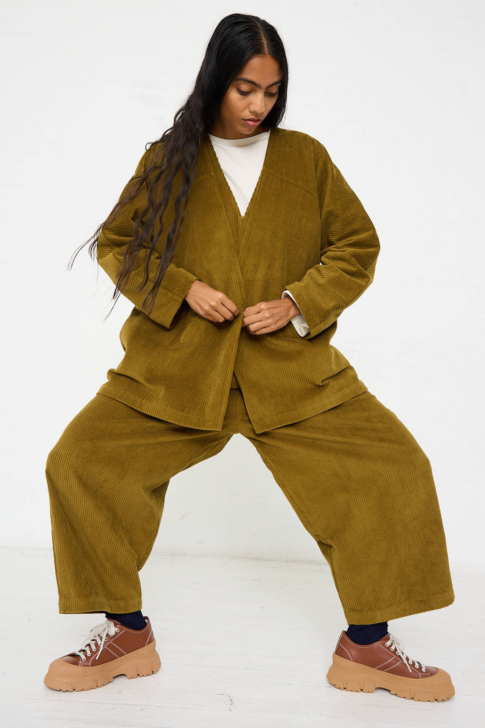 A person with long dark hair wears a loose-fitting Dark Olive Cotton Corduroy Geometric Jacket by Black Crane and brown platform shoes, posing with feet apart and hands adjusting the jacket.