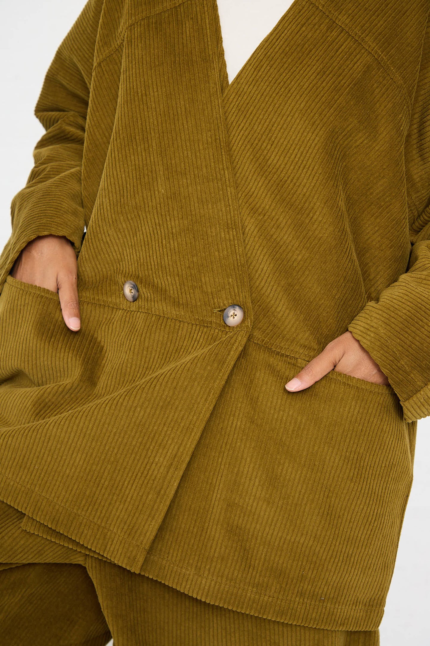 A person wearing a Black Crane Cotton Corduroy Geometric Jacket in Dark Olive with visible buttons and two hands in the pockets.