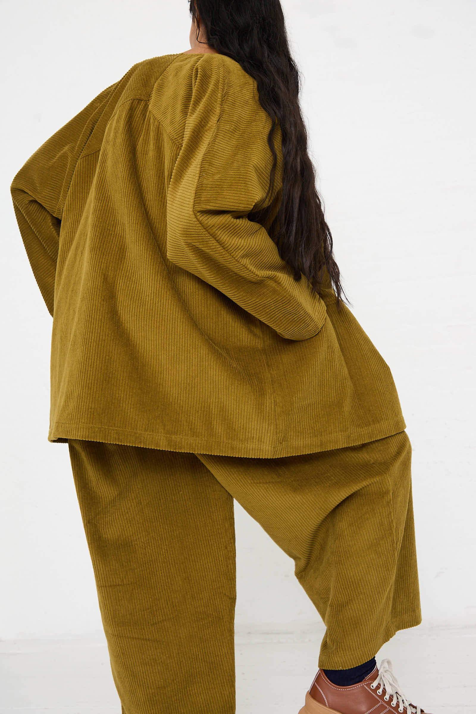 Person with long dark hair wearing a relaxed fit Cotton Corduroy Geometric Jacket in Dark Olive by Black Crane and brown sneakers, posing with their back to the camera against a plain white background.