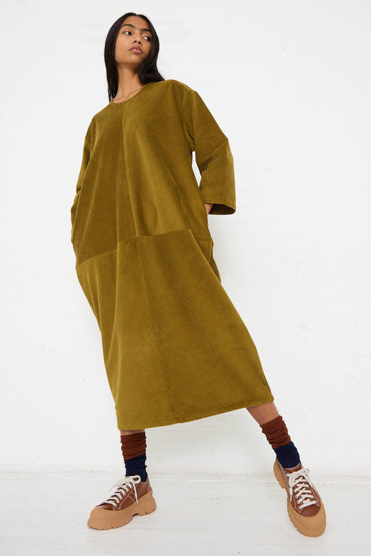 Person standing against a plain white background wearing a dark olive Cotton Corduroy Radient Dress by Black Crane, brown sneakers, and dark socks.