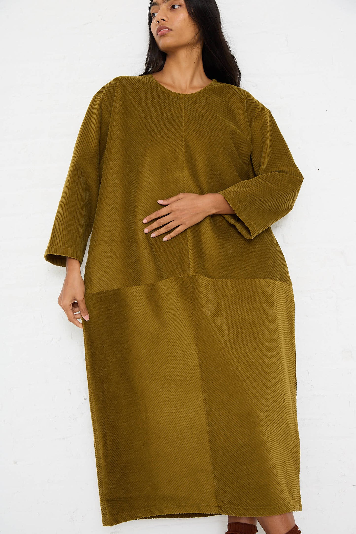 A person with long dark hair wears an oversized, long sleeve Cotton Corduroy Radient Dress in Dark Olive by Black Crane. One hand rests on their stomach as they stand against a plain white background.