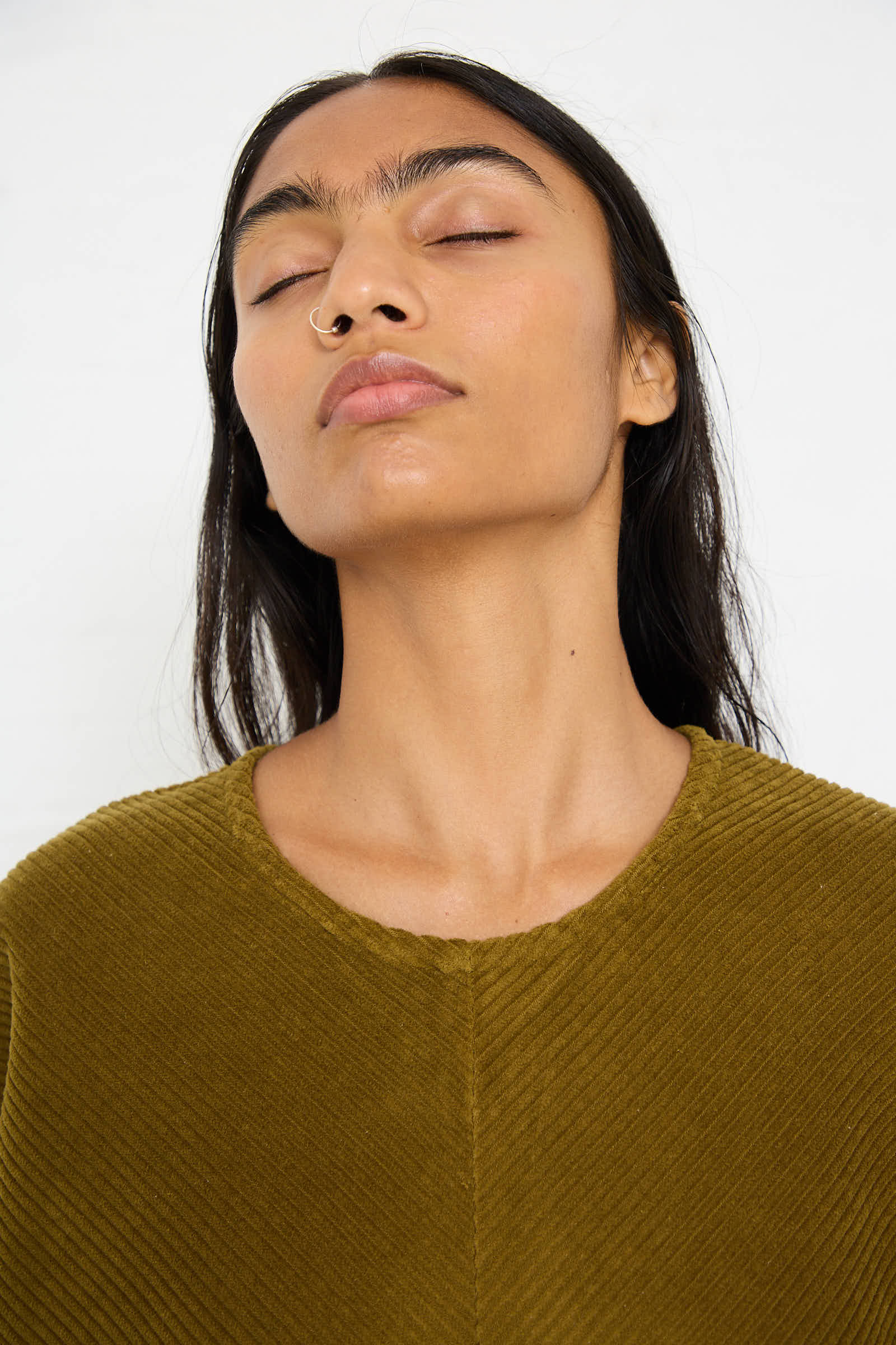 A person with closed eyes and a neutral expression, wearing the Cotton Corduroy Radiant Dress in Dark Olive from Black Crane and a nose ring.