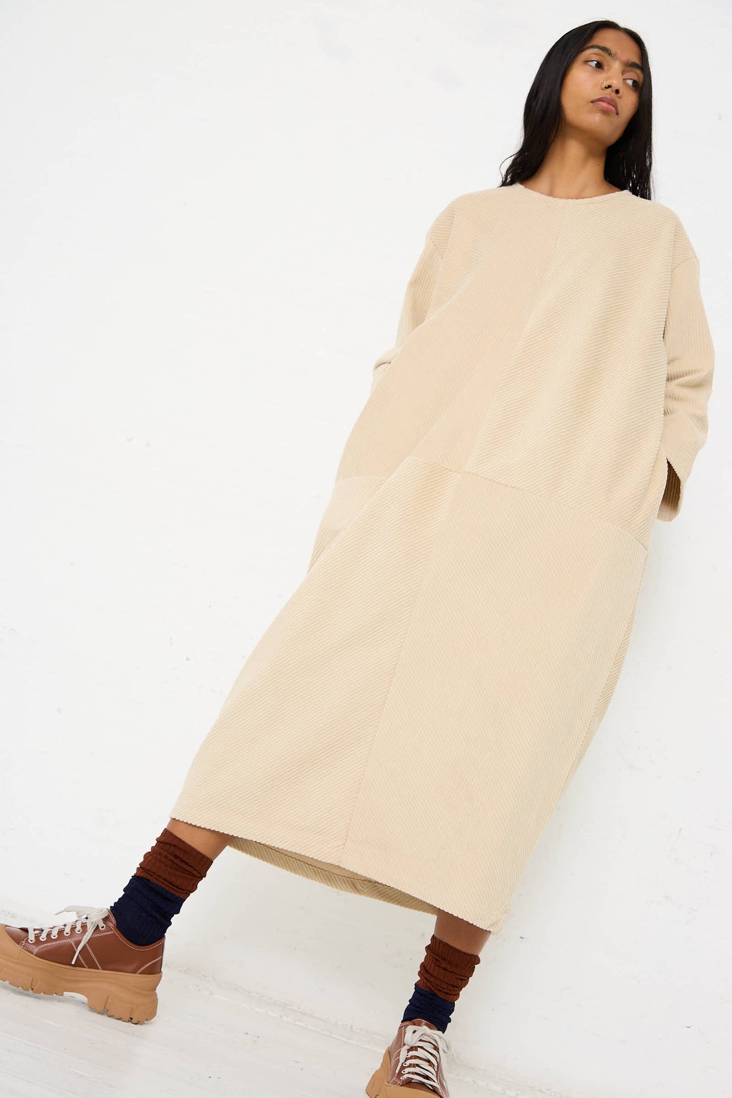 A person stands against a white background wearing the Cotton Corduroy Radiant Dress in Ivory by Black Crane. The long dress, featuring a relaxed fit and pockets where they casually rest their hands, is paired with brown socks layered under black socks and beige lace-up shoes.