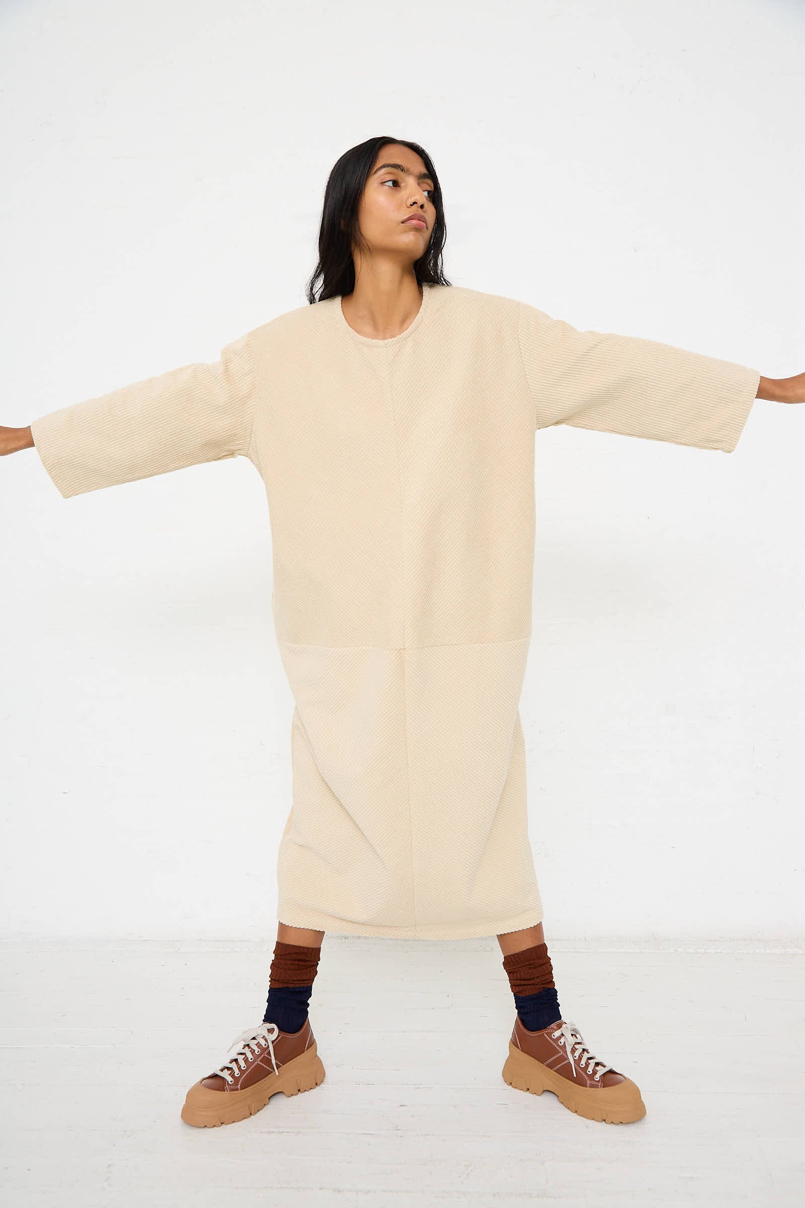 Person standing with arms outstretched, wearing the Black Crane Cotton Corduroy Radiant Dress in Ivory, brown striped socks, and beige platform shoes, against a plain white background.