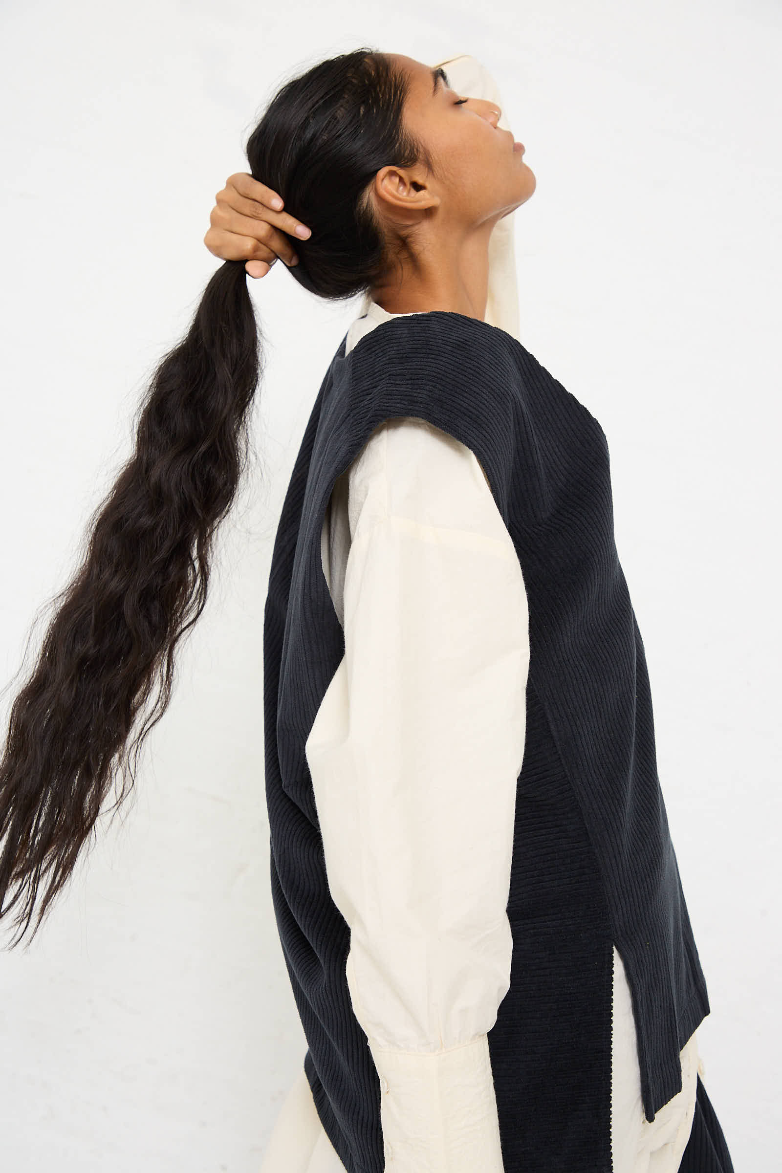 A person holding their long hair in one hand, wearing a Black Crane Cotton Corduroy Vest in Dark Grey over a white long-sleeve shirt, standing against a plain white background.