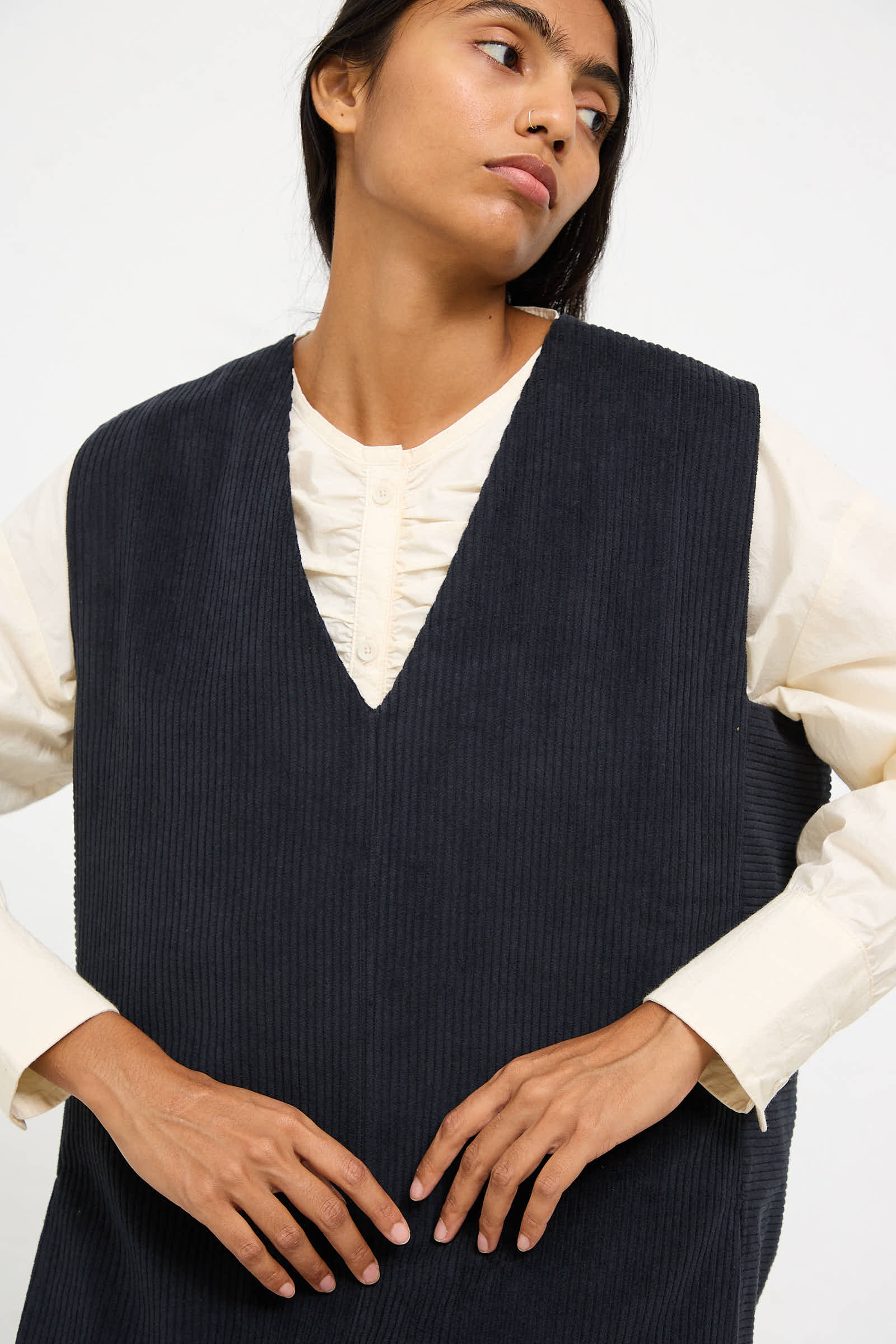 Person wearing a white long-sleeve top under a dark grey Cotton Corduroy Vest from Black Crane, looking slightly to the side with one hand resting on the vest.