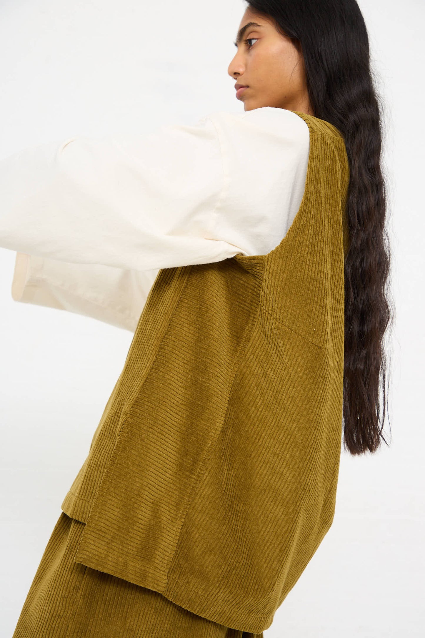 Person with long dark hair wears a white long-sleeved shirt under a Black Crane Cotton Corduroy Vest in Dark Olive, seen from the side against a plain background.