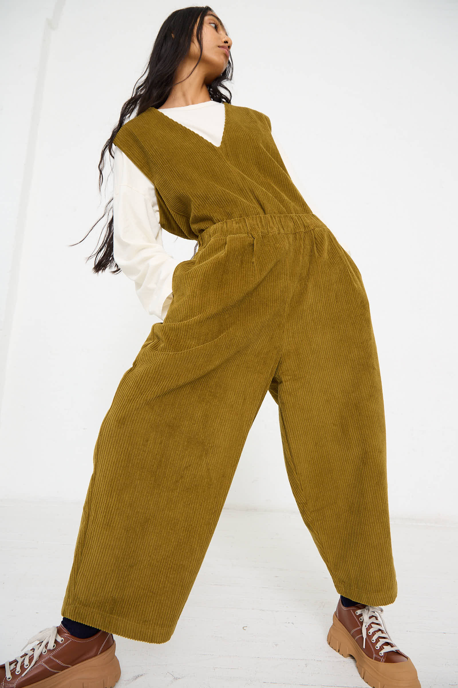 A person stands against a white background, looking upward with hair flowing down their back, wearing the Black Crane Cotton Corduroy Wide Pant in Dark Olive and brown sneakers.