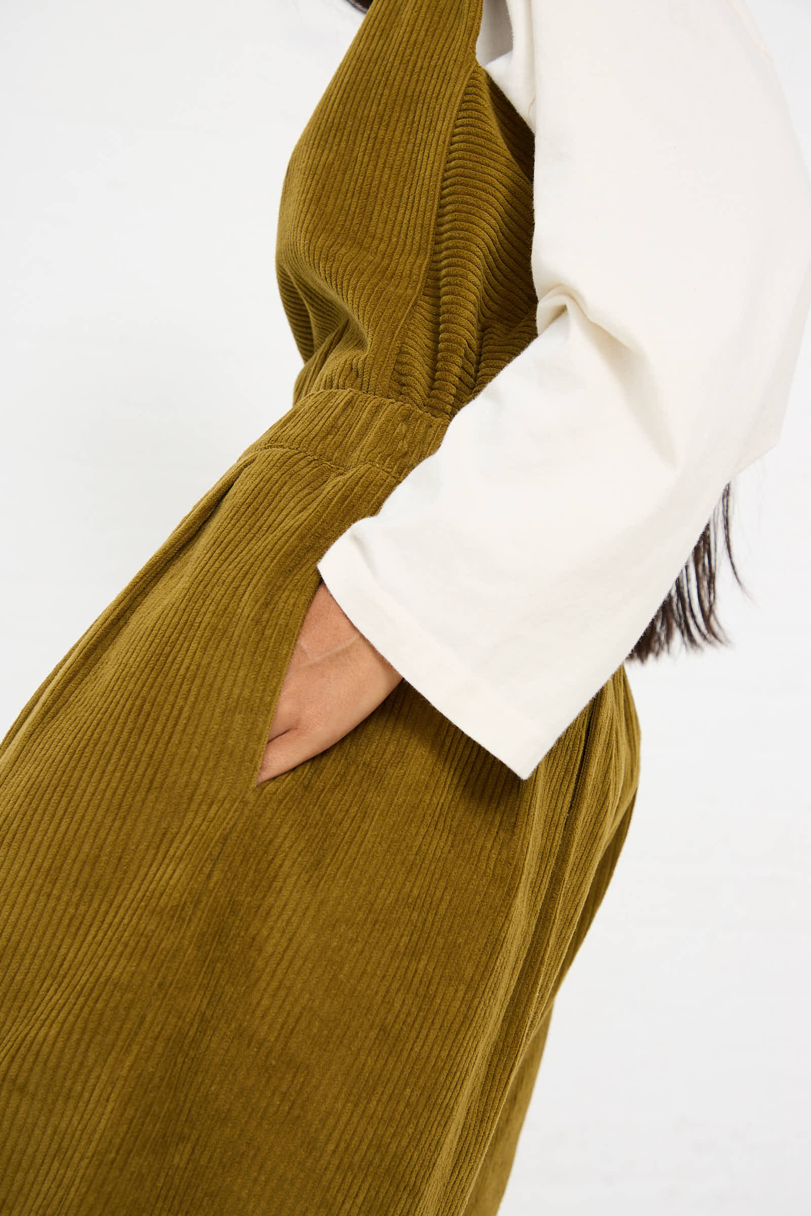 A person wearing a white long-sleeve shirt and an olive green corduroy dress with a hand in the dress pocket, paired seamlessly with Black Crane's Cotton Corduroy Wide Pant in Dark Olive.