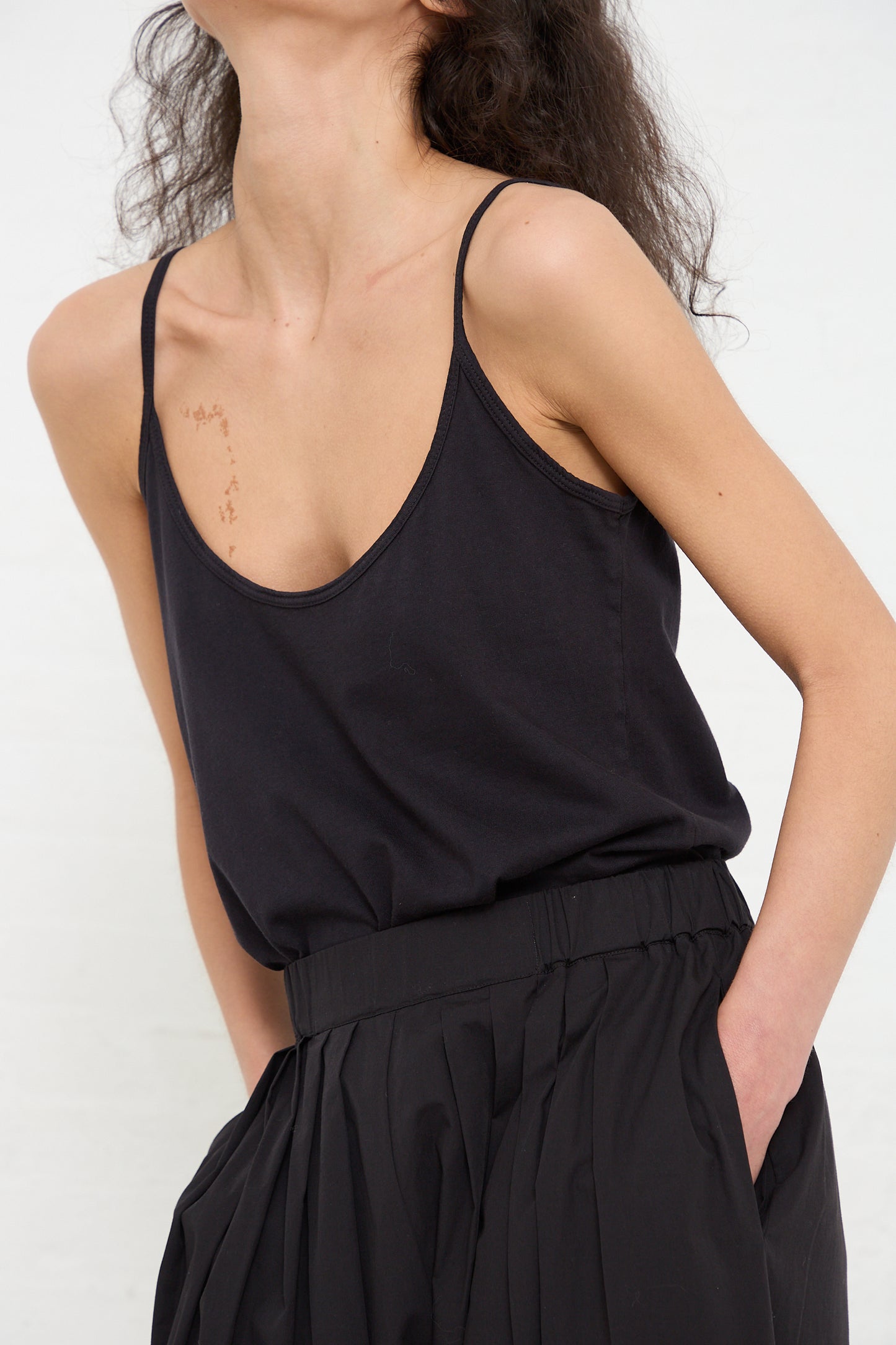 A person in a Black Crane black organic cotton jersey camisole top and pleated skirt poses with one hand on hip, showcasing the outfit against a plain background.