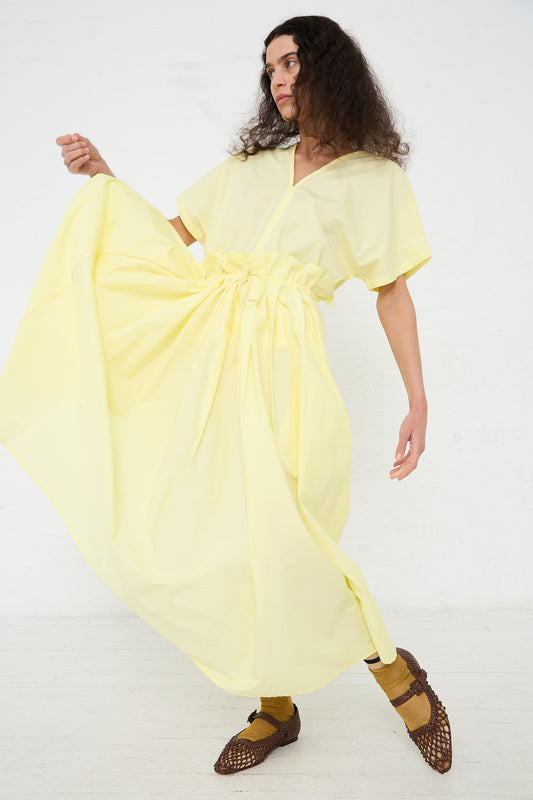 Woman posing in a yellow dress with puff sleeves and a knotted detail, paired with brown woven shoes and a Black Crane Organic Cotton Parachute Skirt in Lemon.