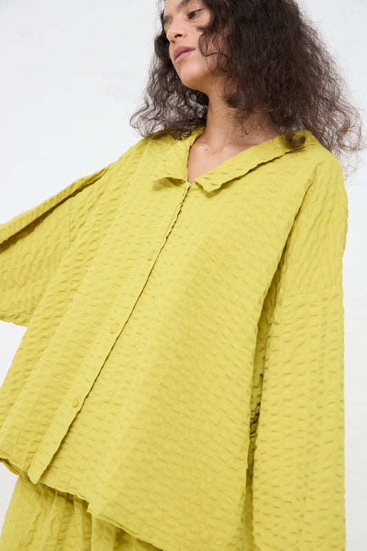A woman in a Cotton Seersucker Chelsea Collar Shirt in Turmeric by Black Crane, with her head tilted slightly downwards, against a white background.