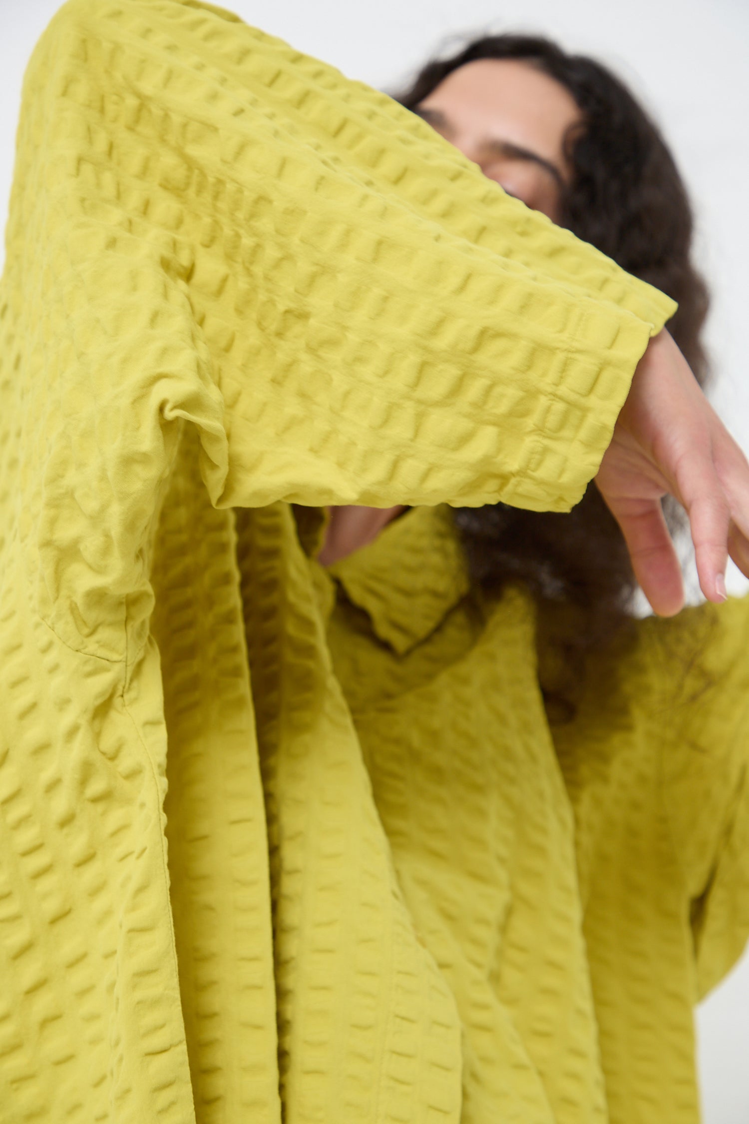 A person in a yellow Cotton Seersucker Chelsea Collar Shirt in Turmeric by Black Crane, with their arm raised partially obscuring their face.