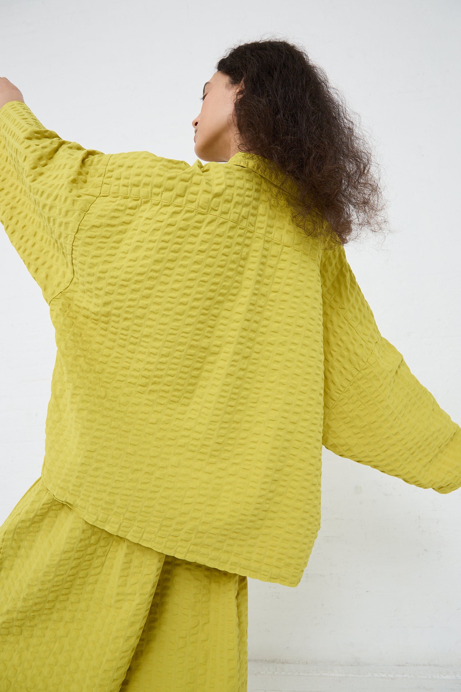 A woman in a Cotton Seersucker Chelsea Collar Shirt in Turmeric by Black Crane, with her back to the camera, looking to one side with her left arm extended outwards.