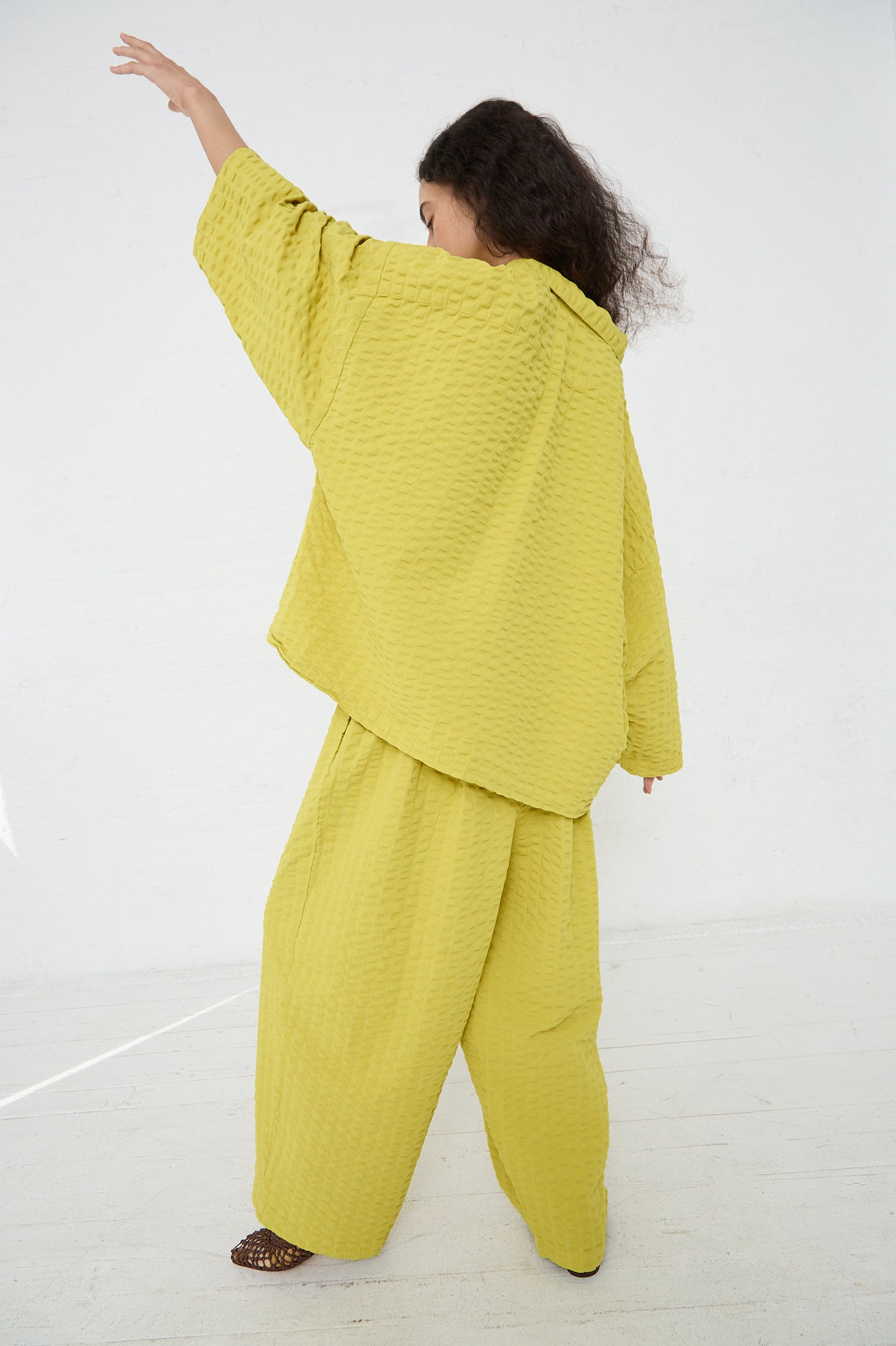 A woman in a Yellow Cotton Seersucker Chelsea Collar Shirt in Turmeric by Black Crane is standing with her back to the camera, extending her right arm upwards. Her left leg is slightly raised, visible from a side