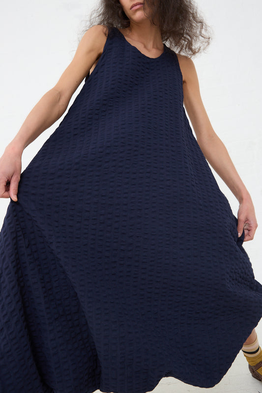 A person wearing a textured navy blue sleeveless Black Crane cotton seersucker parachute dress, holding the fabric out to the sides against a white background.