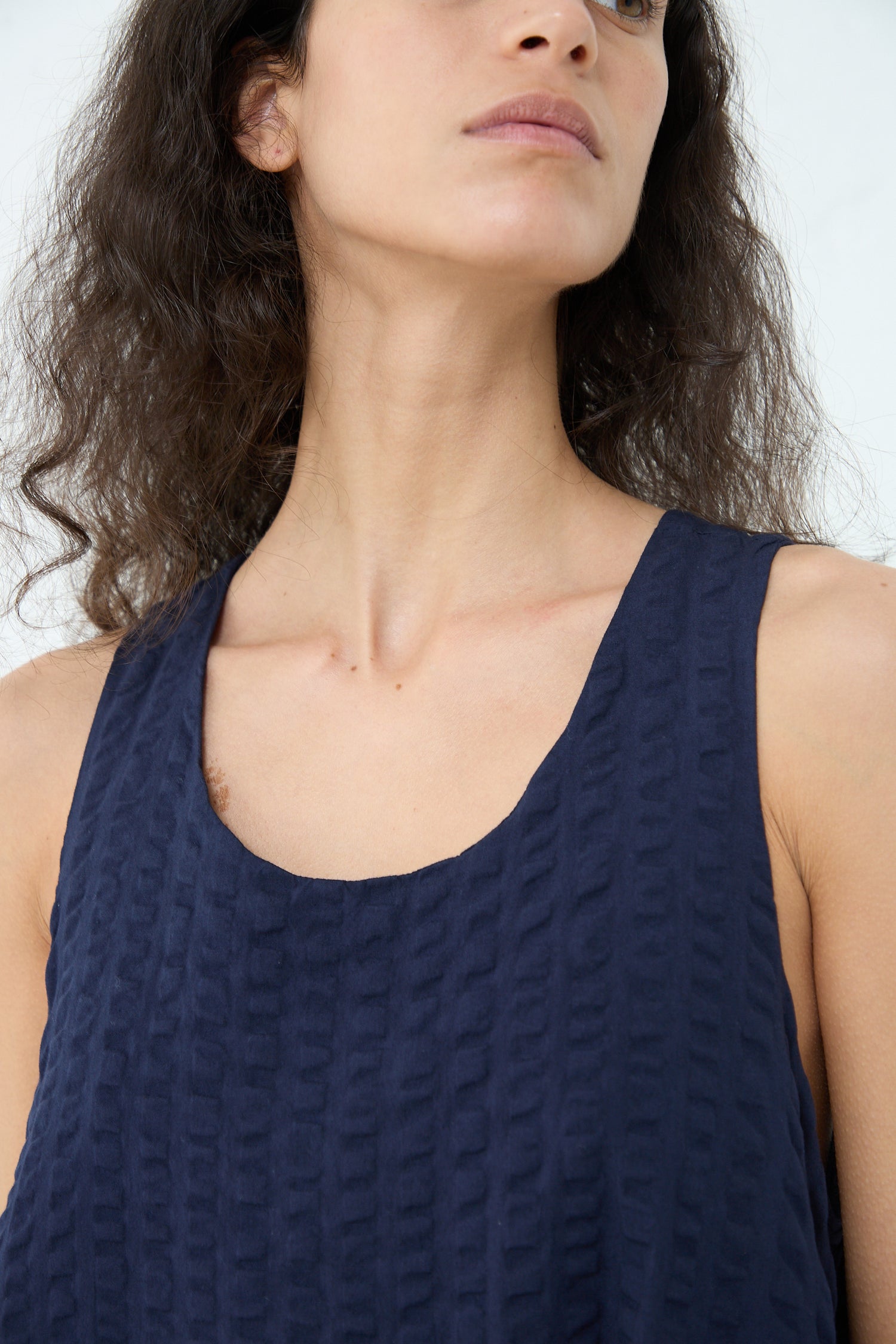 A woman wearing a Black Crane Cotton Seersucker Parachute Dress in Navy, showing her shoulders and collarbone area, with her face partially visible and her hair falling naturally.