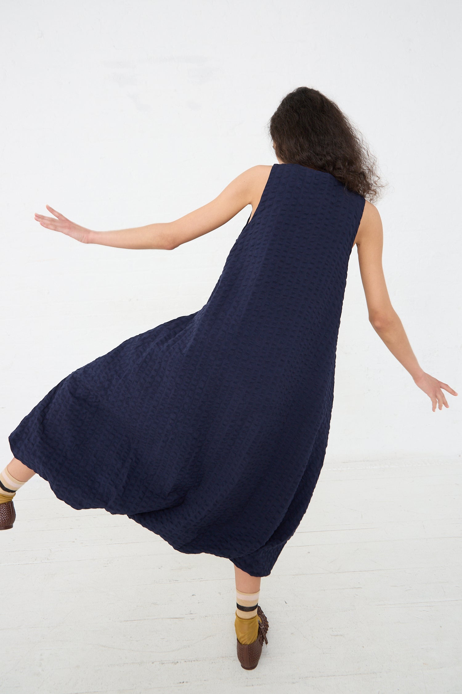 A woman with curly hair twirls in a spacious room while wearing a Navy Cotton Seersucker Parachute Dress from Black Crane and brown shoes.
