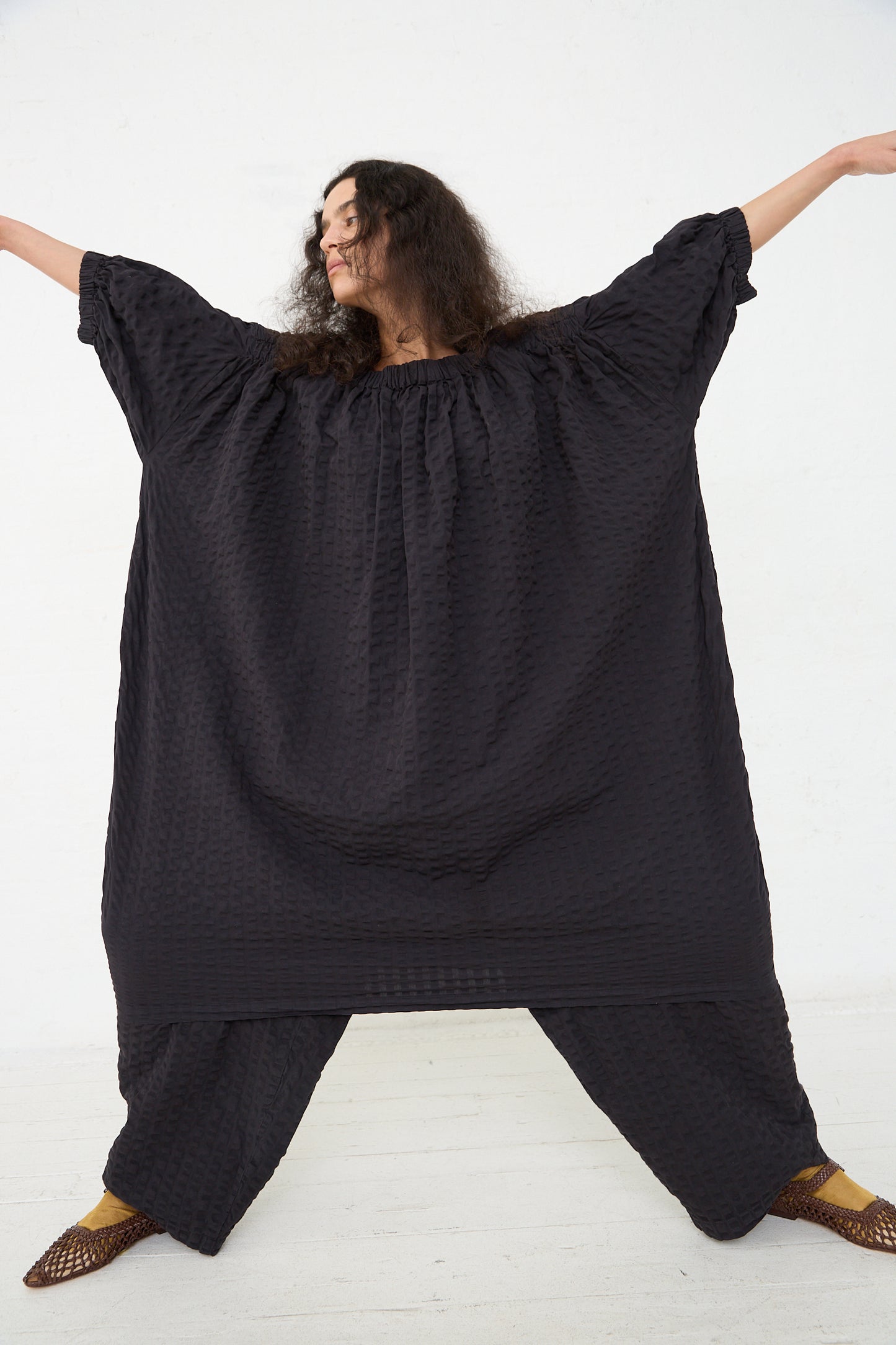 A person wearing an oversized Black Crane Cotton Seersucker Sack Dress in Ink Black with a ruched neckline and wide sleeves stands against a white background, arms outstretched to display the full breadth of the outfit.