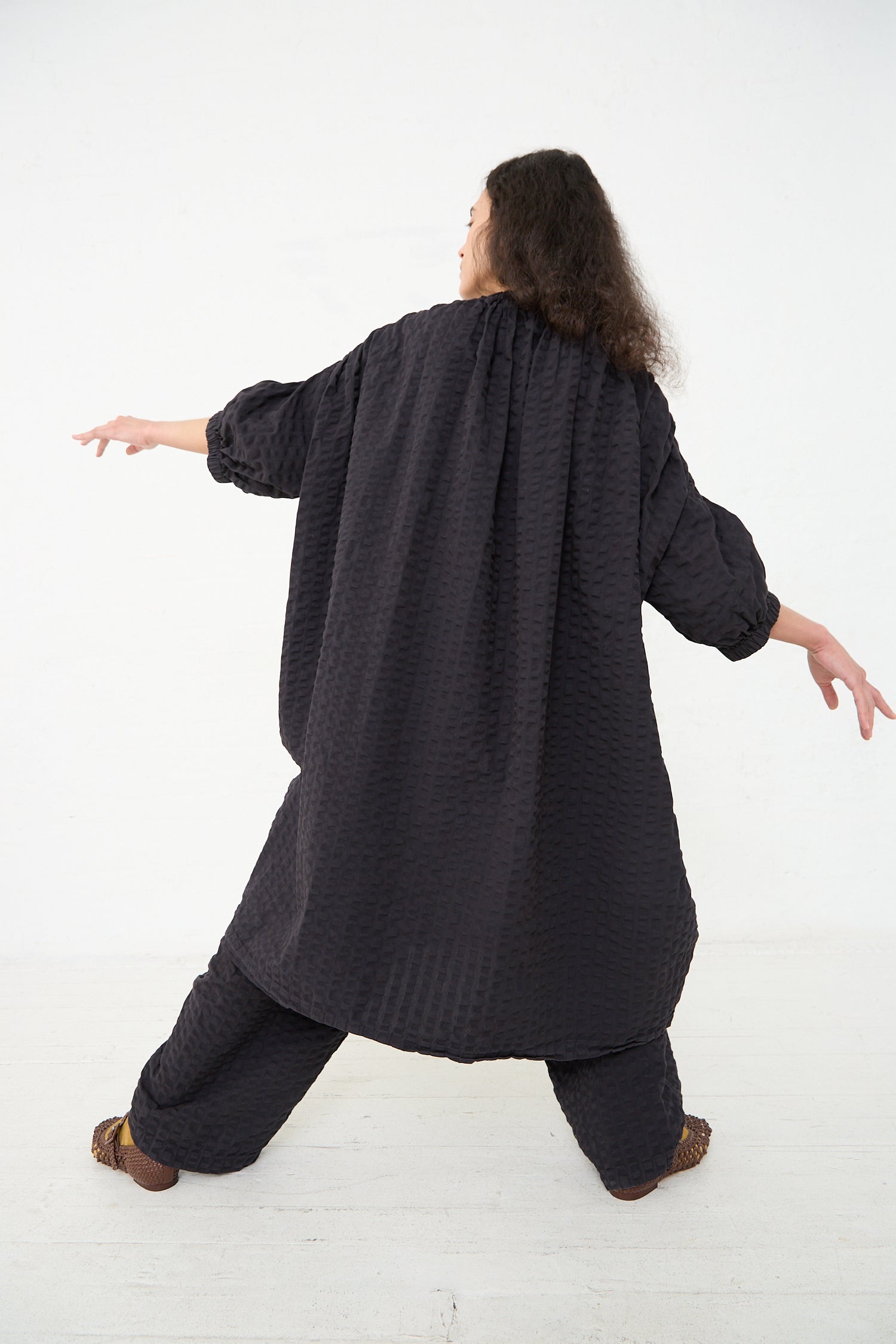 A person wearing a Black Crane Cotton Seersucker Sack Dress in Ink Black stands with their back to the camera, arms extended outward in a t-pose, and one leg stepping forward, against a plain white background.