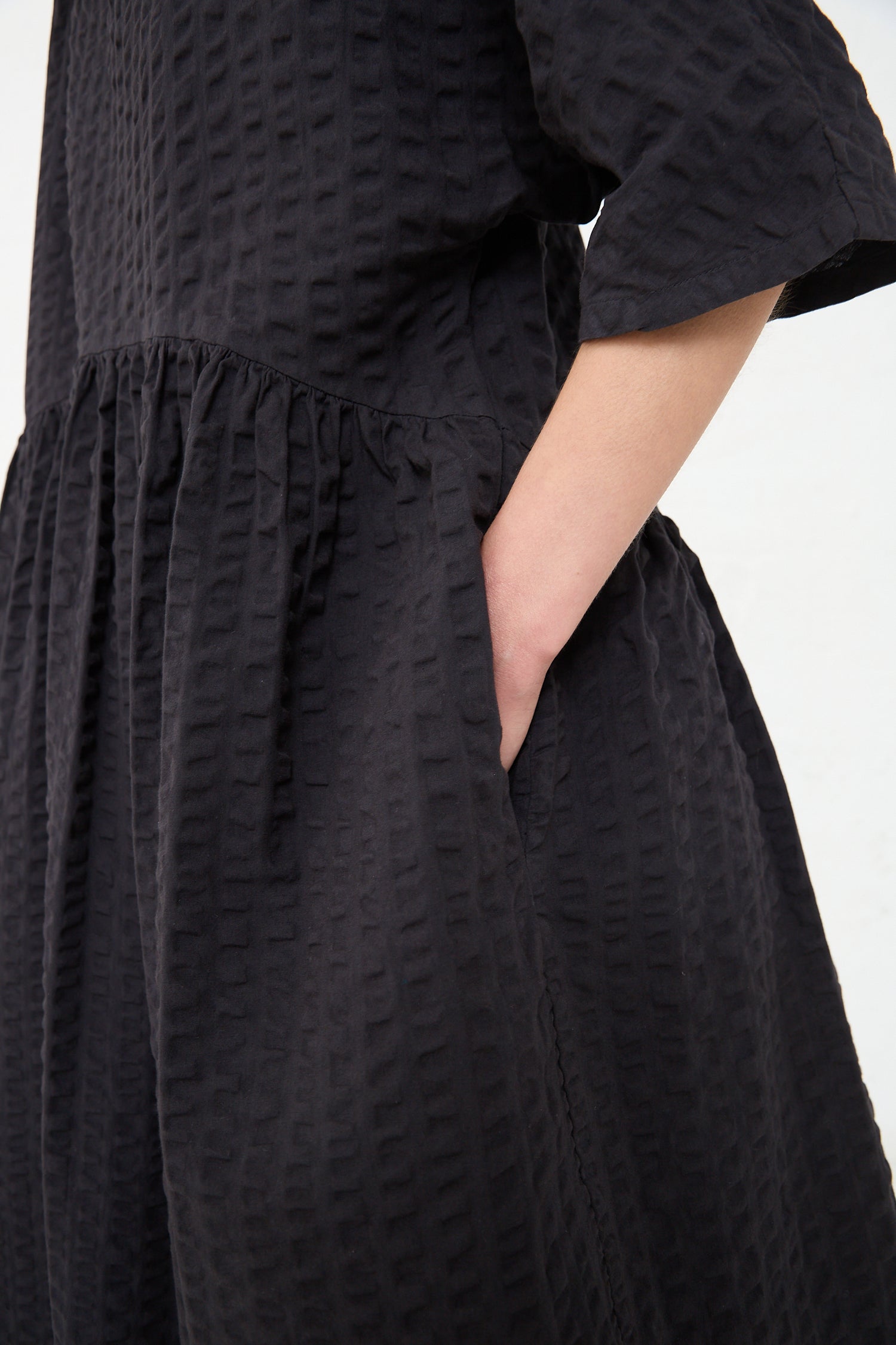 A person is wearing a Cotton Seersucker Tradi Dress in Ink Black by Black Crane with a cinched waist detail. Only the torso and part of the arm are visible in the frame.