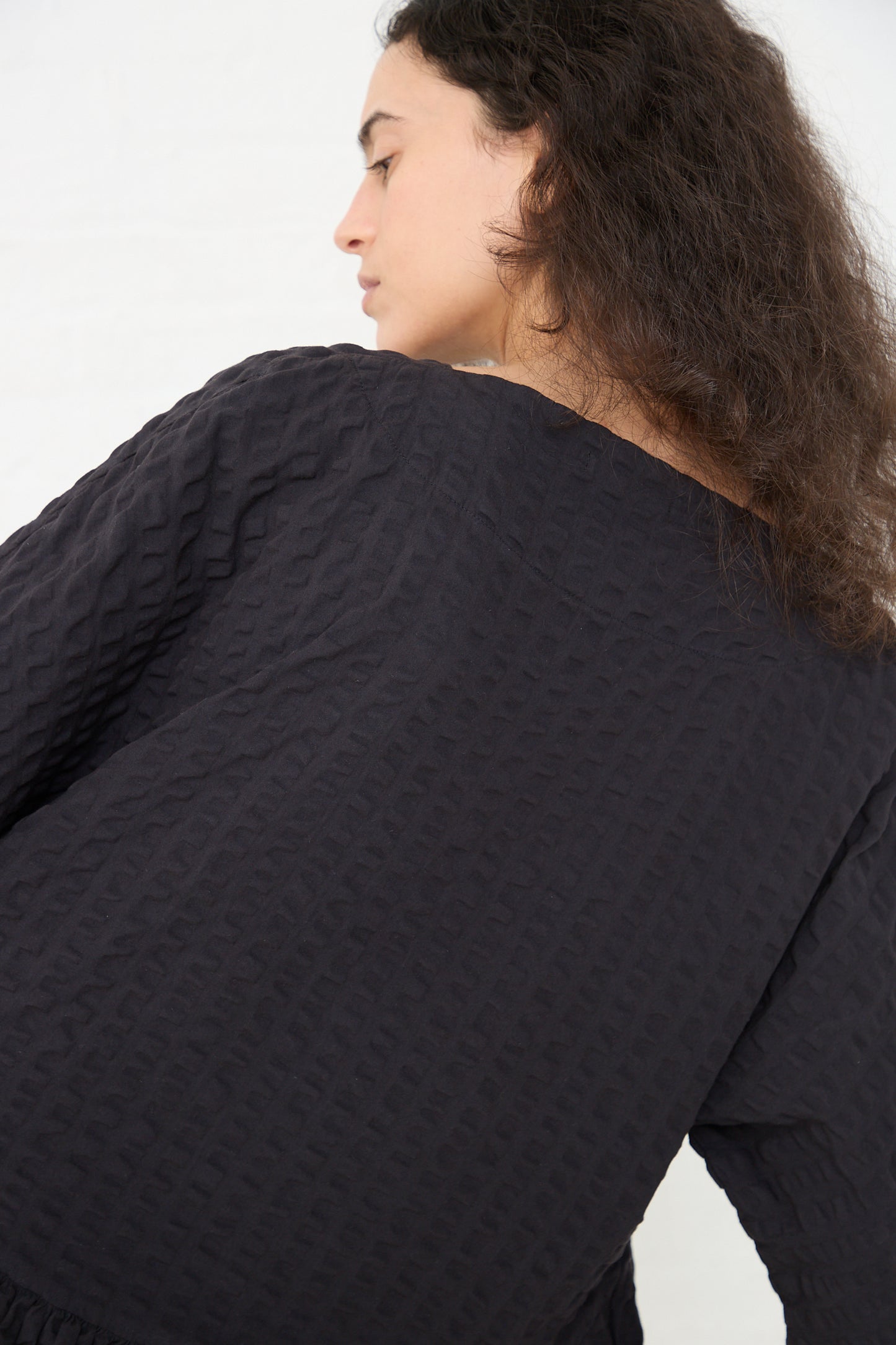 Profile view of a woman with long curly hair wearing a textured Black Crane Cotton Seersucker Tradi Dress in Ink Black top.
