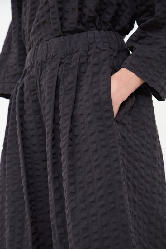 A person is pictured wearing textured black cotton seersucker wide leg pants from Black Crane with their hand resting on their waist.