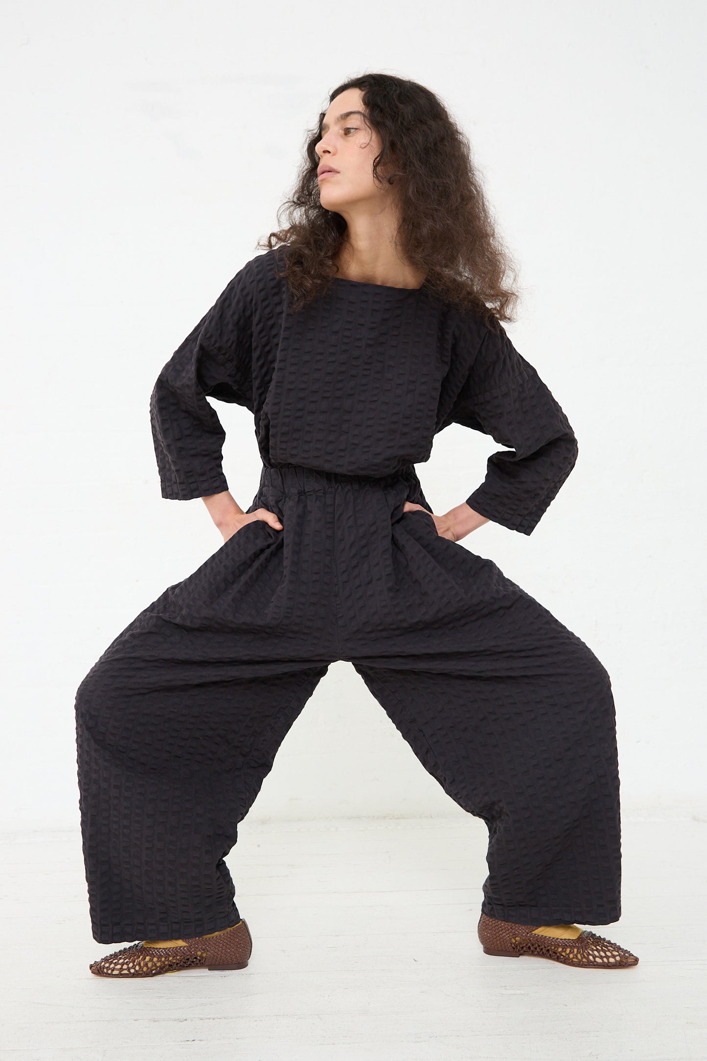 A person stands with hands on hips, wearing a textured Black Crane Cotton Seersucker Wide Pant in Ink Black jumpsuit and patterned flat shoes against a plain white background.