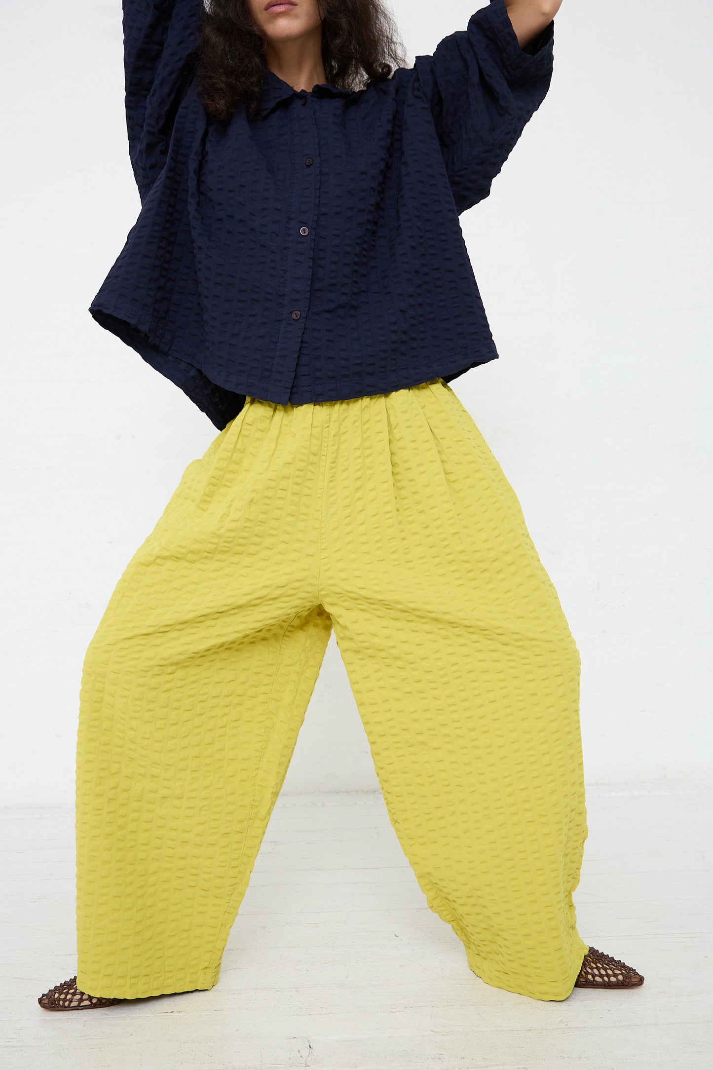 A person stands against a white background wearing a navy blue blouse with voluminous sleeves and high-waisted Yellow Cotton Seersucker Wide Pant in Turmeric trousers by Black Crane, with brown flat shoes visible.