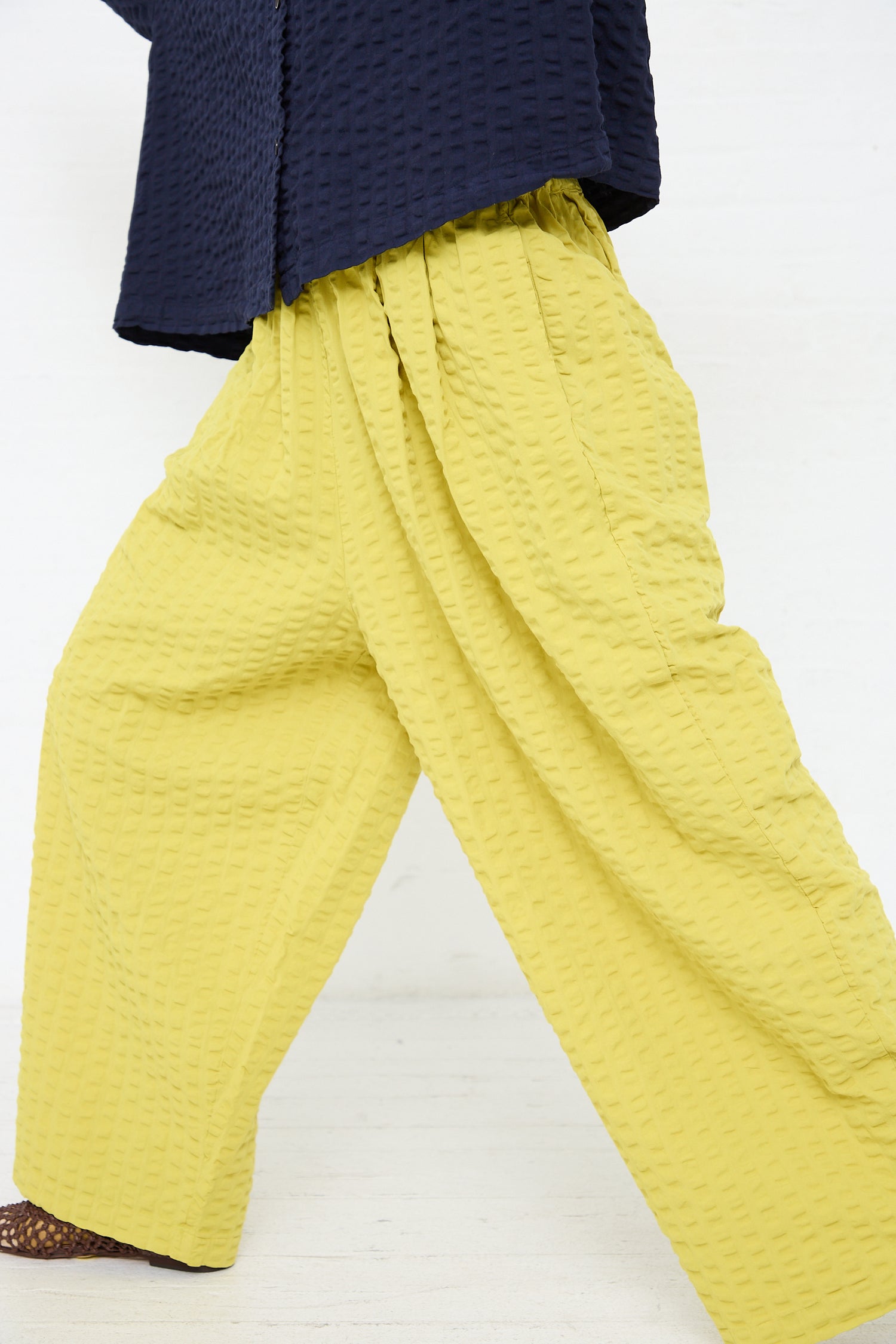 A person standing with their back to the camera, wearing a Cotton Seersucker Wide Pant in Turmeric by Black Crane and a portion of a dark blue quilted jacket visible, against a white background.