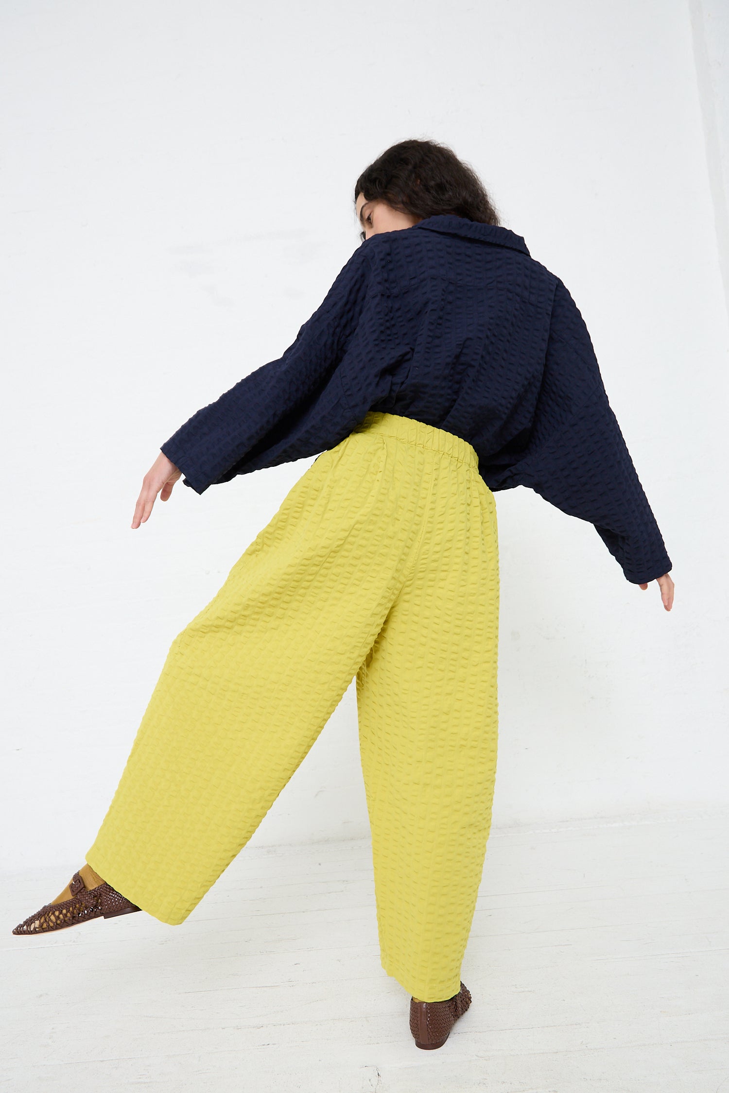 A person wearing a navy blue textured top and Black Crane's Cotton Seersucker Wide Pant in Turmeric with brown patterned shoes, striking a pose with their back to the camera in a white room.
