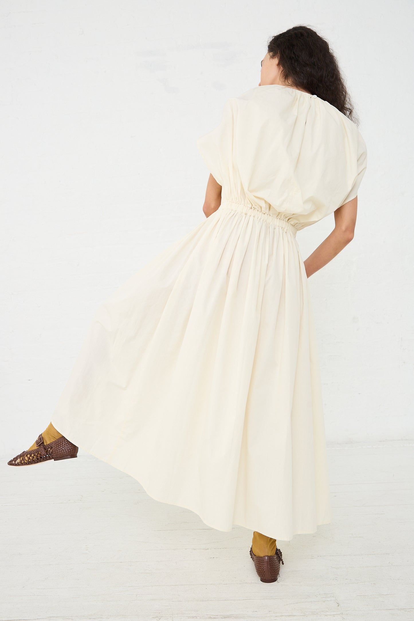 Woman in a sleeveless, flowing Black Crane Organic Cotton Shell Dress in Cream with a cinched waist, posing from the back.