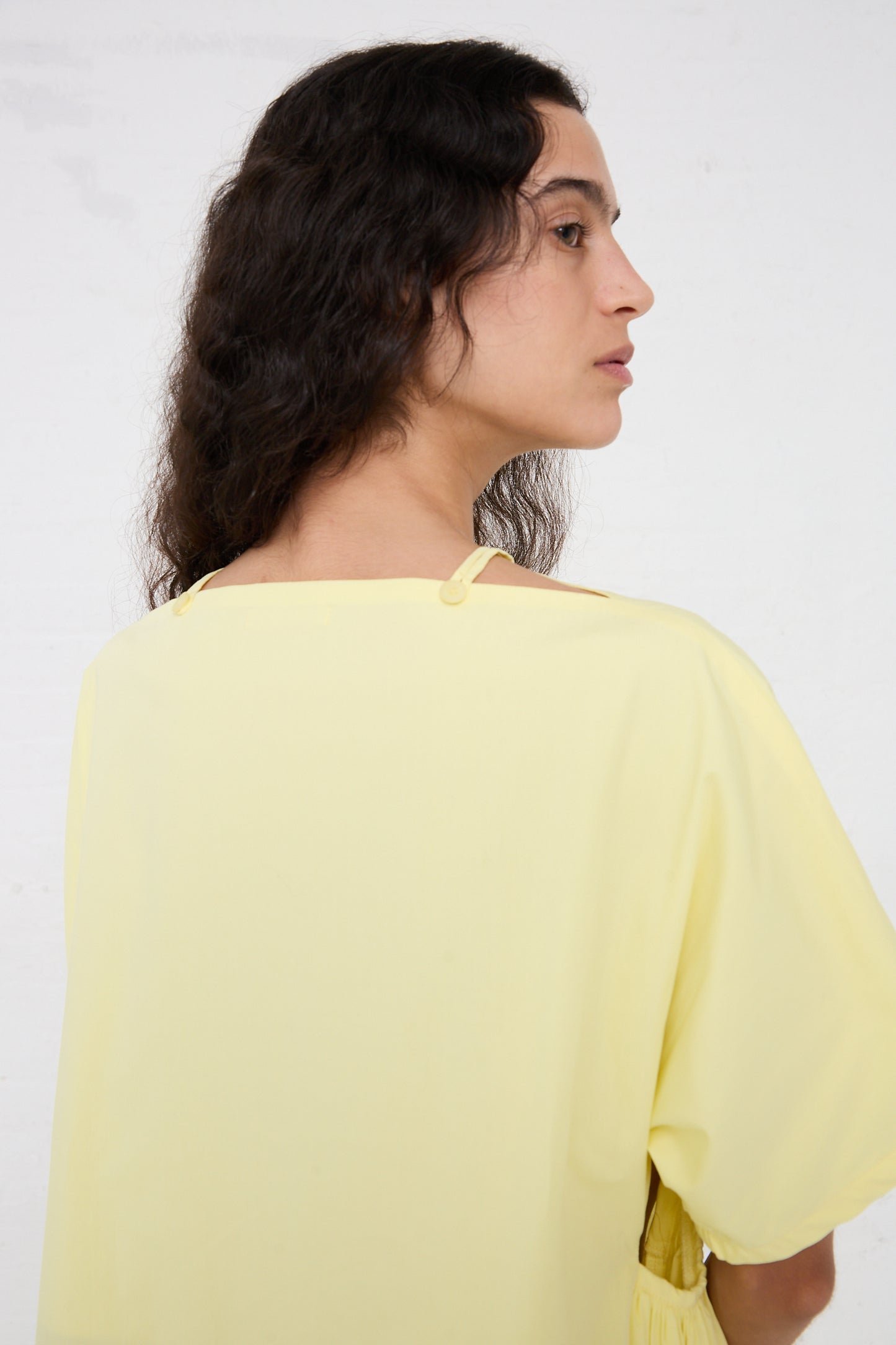 A side profile of a person with shoulder-length curly hair, wearing a Black Crane Organic Cotton Star Neck Dress in Lemon, featuring a buttoned back detail.