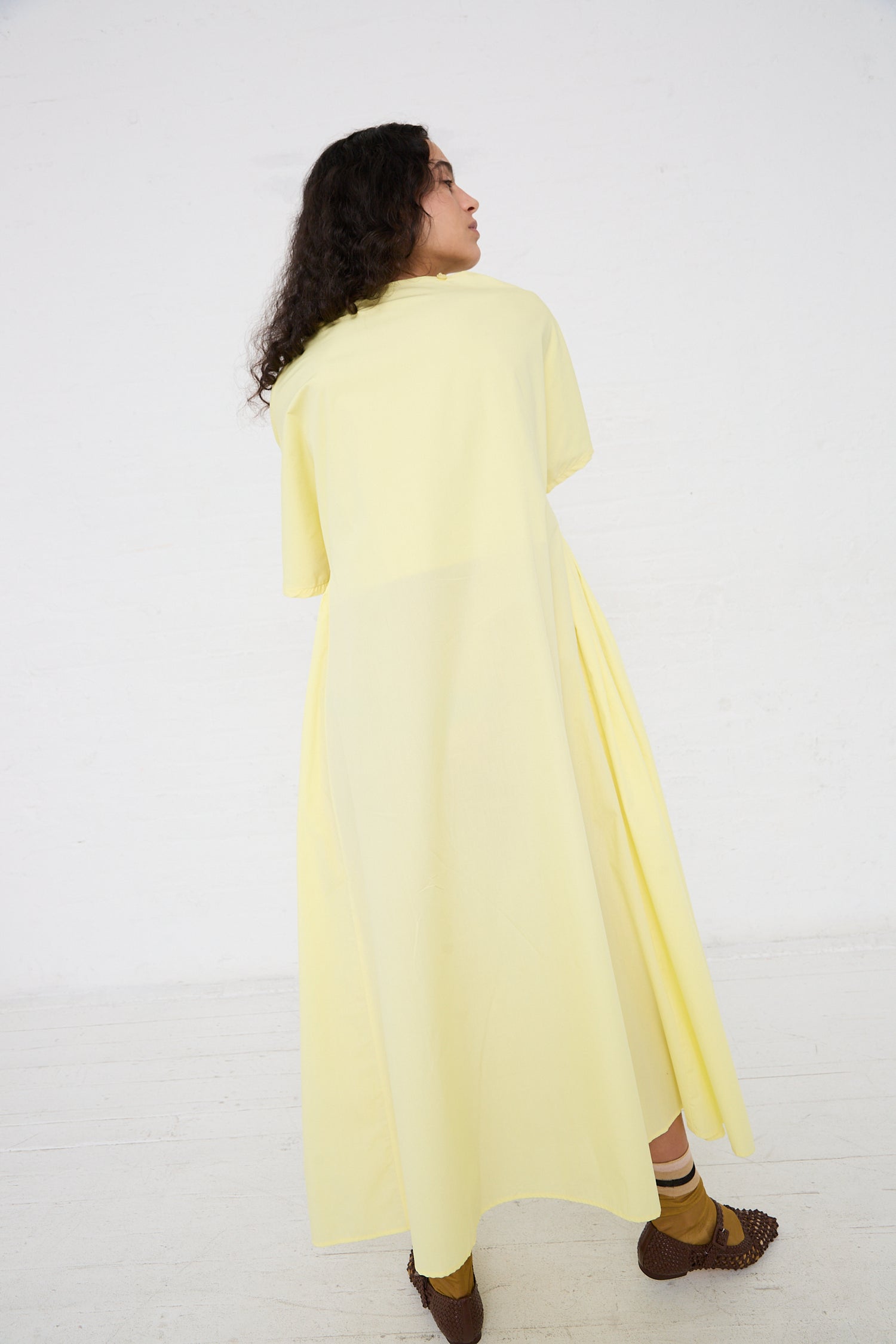 Woman in a yellow Black Crane Organic Cotton Star Neck Dress in Lemon looking over her shoulder against a white background.