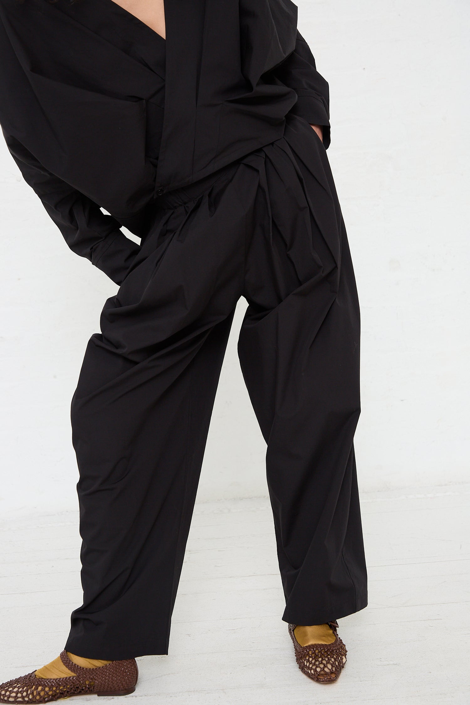 A person wearing Black Crane's Organic Cotton Straight Draped Pant in Black and a matching jacket, paired with brown patterned shoes.