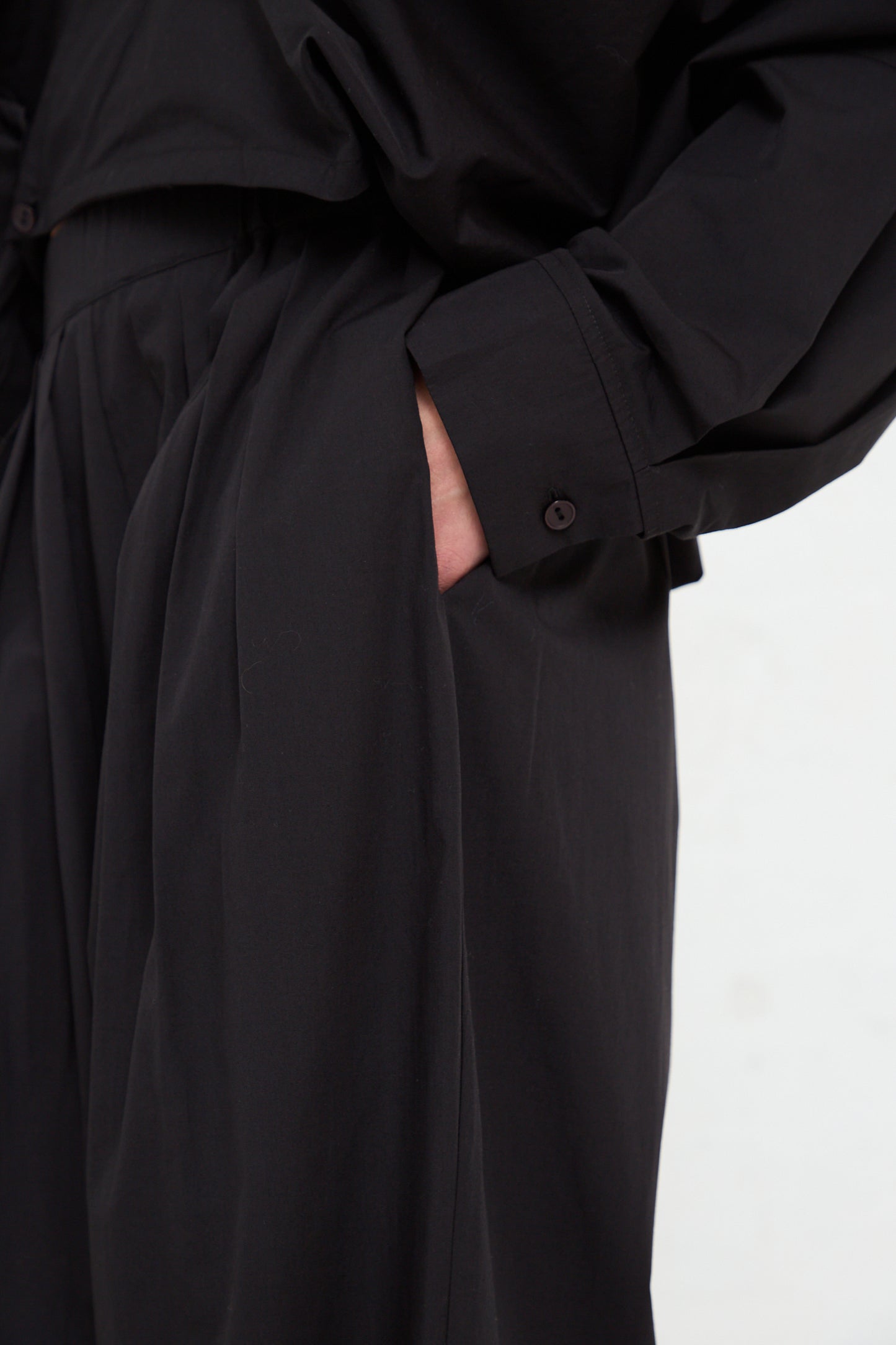 Close-up of a person wearing a Black Crane black trench coat and Black Crane organic cotton straight draped pant in black, with their hand in their pocket.