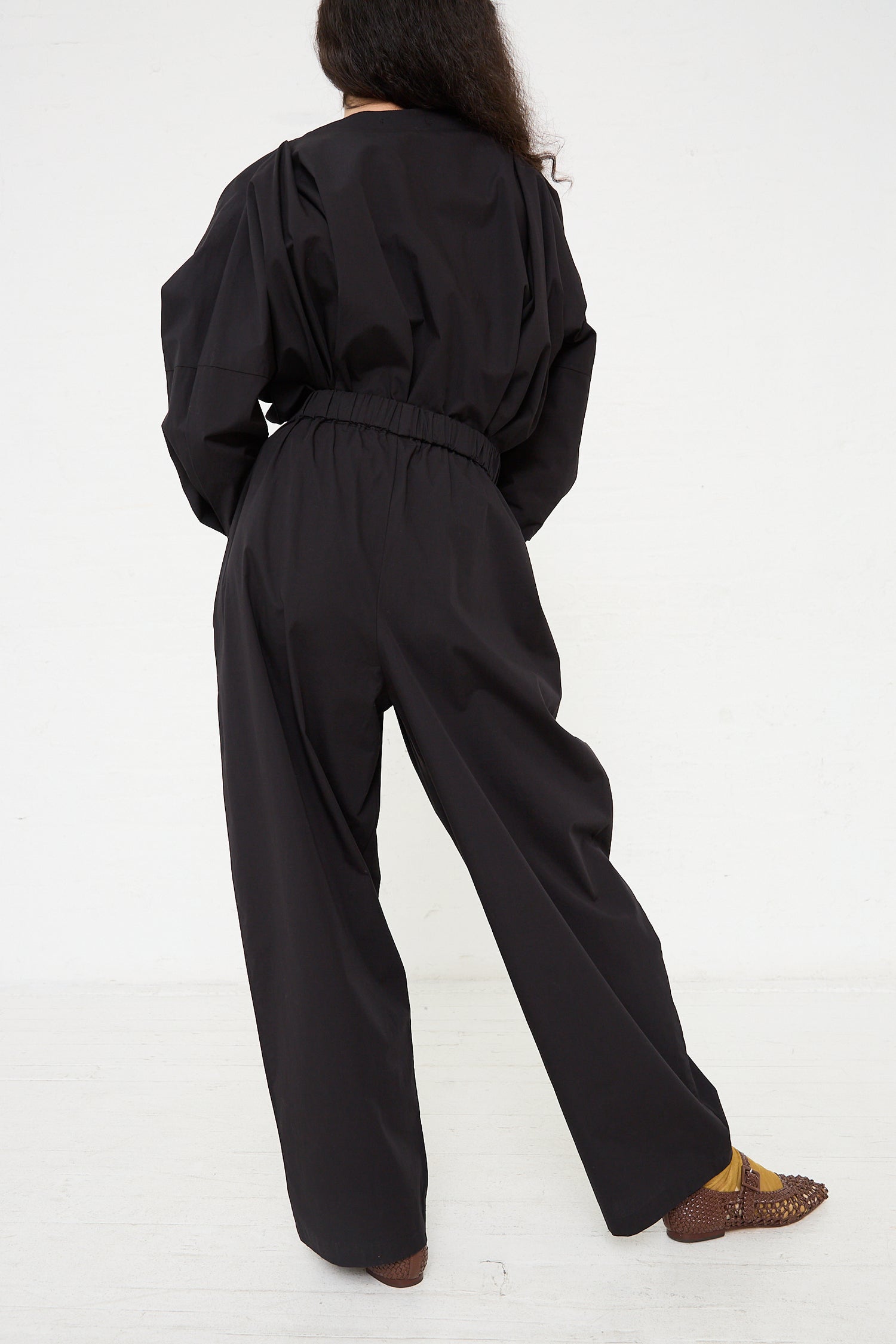 Woman wearing a Black Crane Organic Cotton Straight Draped Pant in Black with gathered waist from behind, paired with patterned shoes.