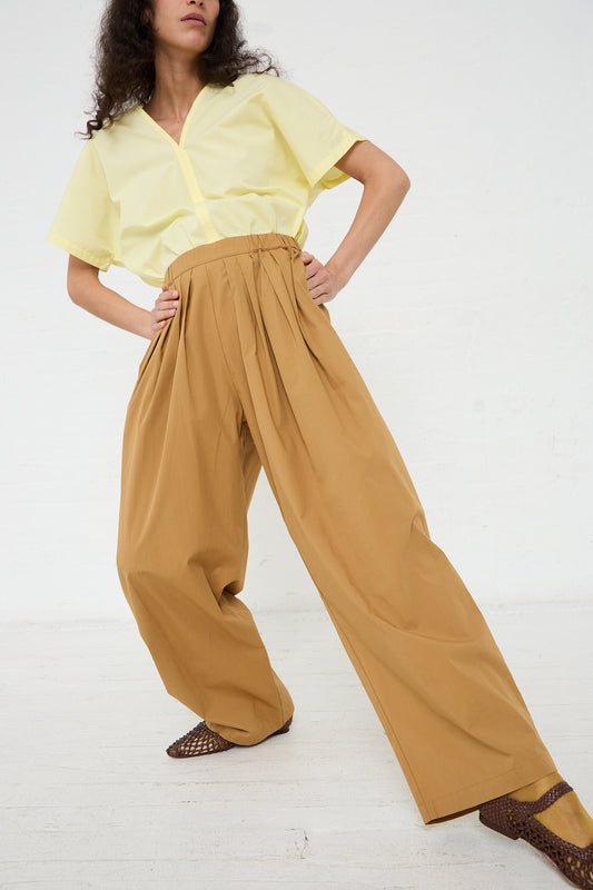 A person posing in a yellow shirt and Black Crane Organic Cotton Straight Draped Pant in Camel with hands in pockets.