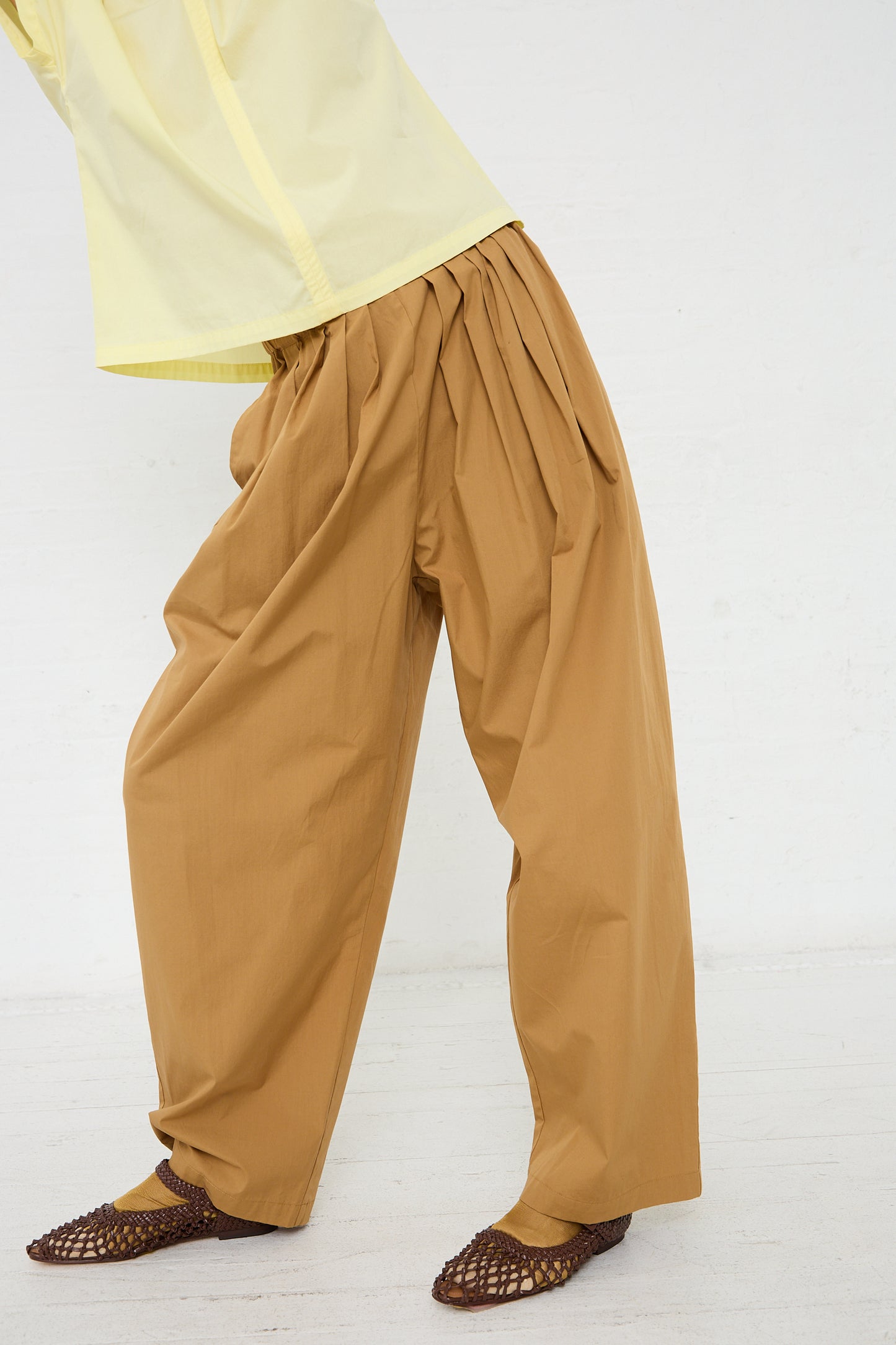 A person standing profile wearing brown straight leg pants and brown woven shoes with the hem of a yellow Black Crane Organic Cotton Straight Draped Pant in Camel visible at the top.