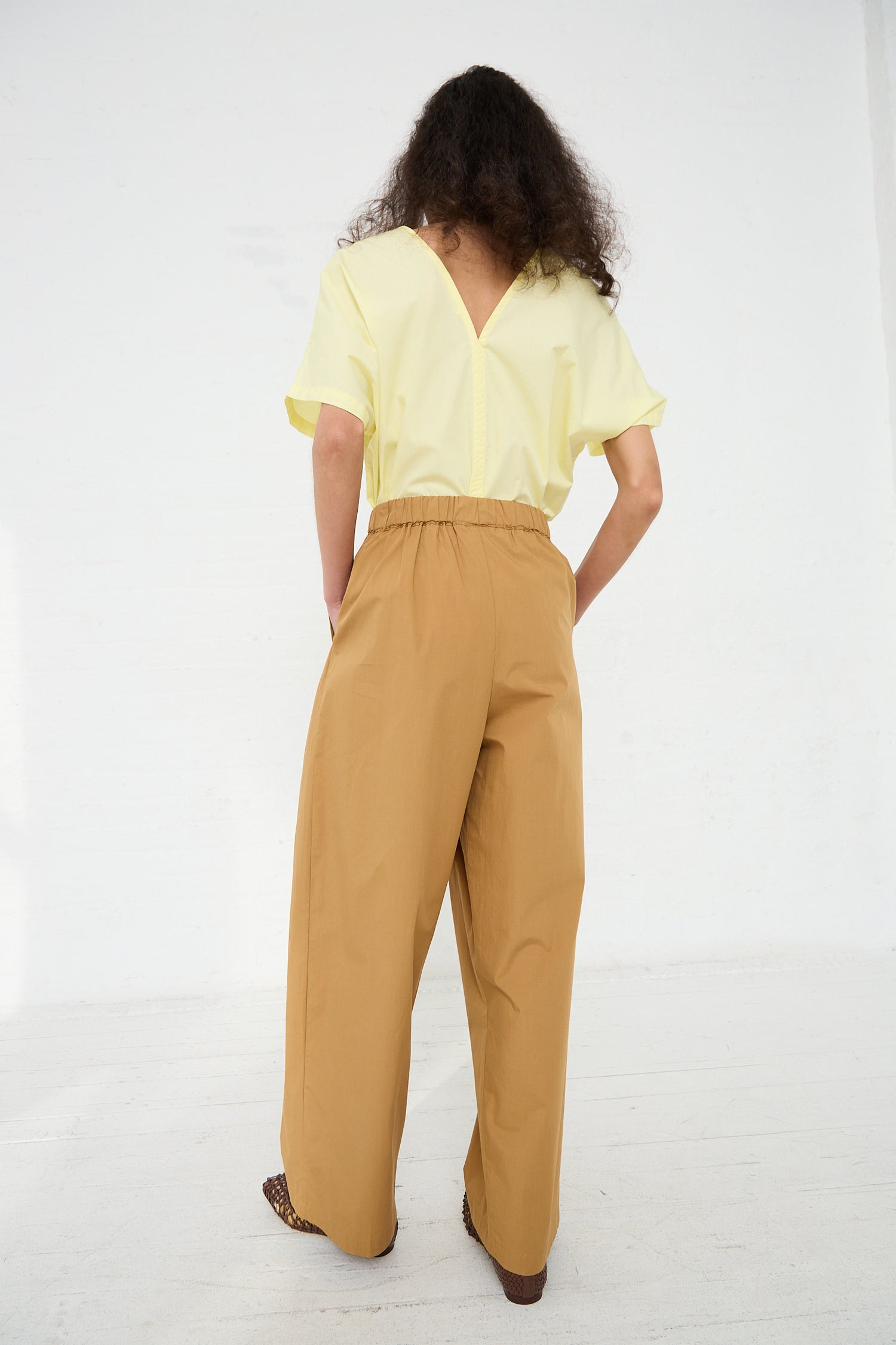 Woman from behind wearing yellow blouse and Black Crane's Organic Cotton Straight Draped Pant in Camel, standing against a white background.