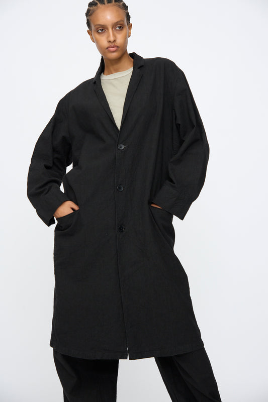 A person stands with hands in pockets, wearing the Black Crane Crinkled Coat in Black—a long Japanese linen coat with a subtle crinkled texture—over a light shirt and black pants, set against a plain white background.
