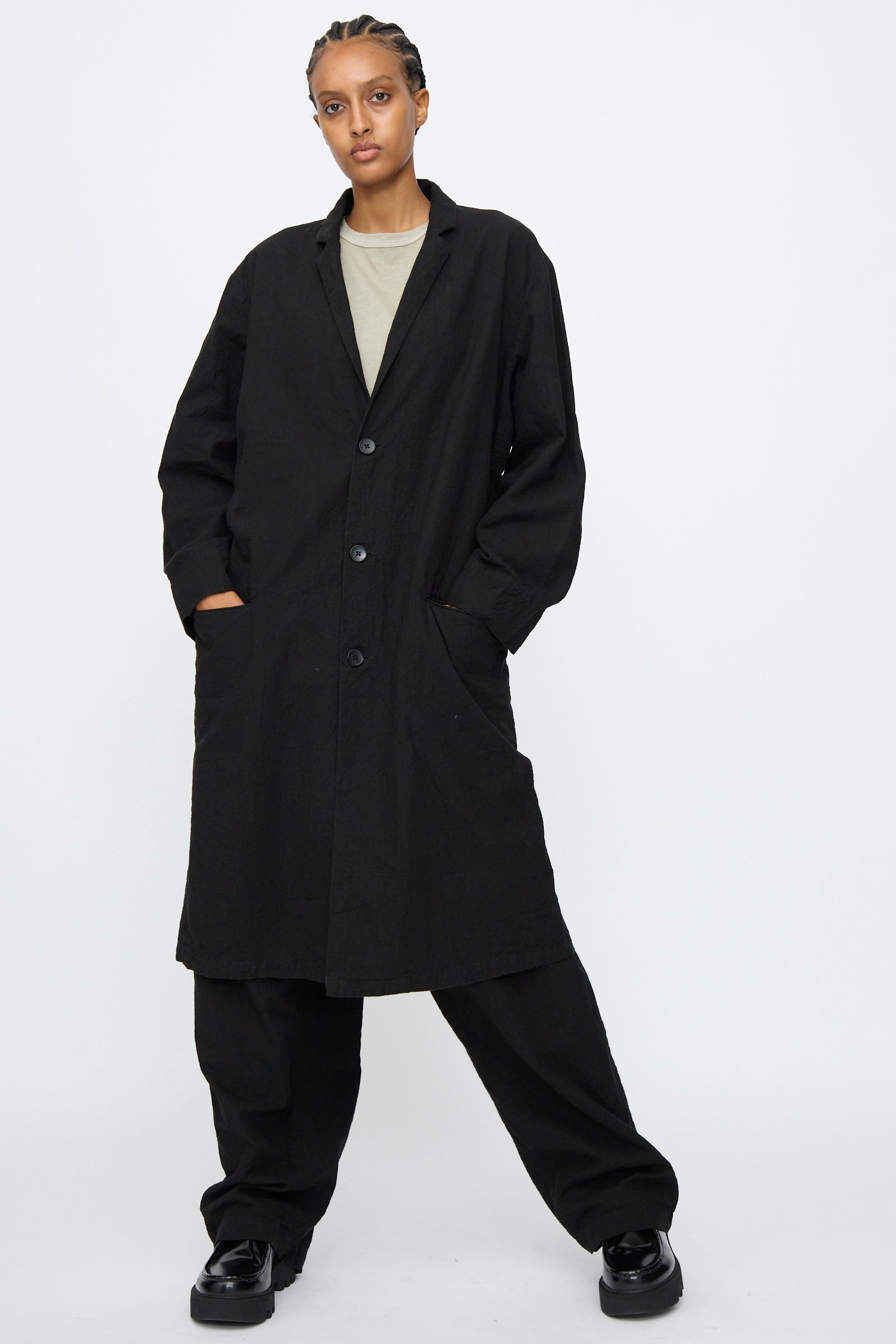 A person wears the Black Crane Crinkled Coat in Black over black pants and a beige shirt, standing with hands in pockets against a plain white background.