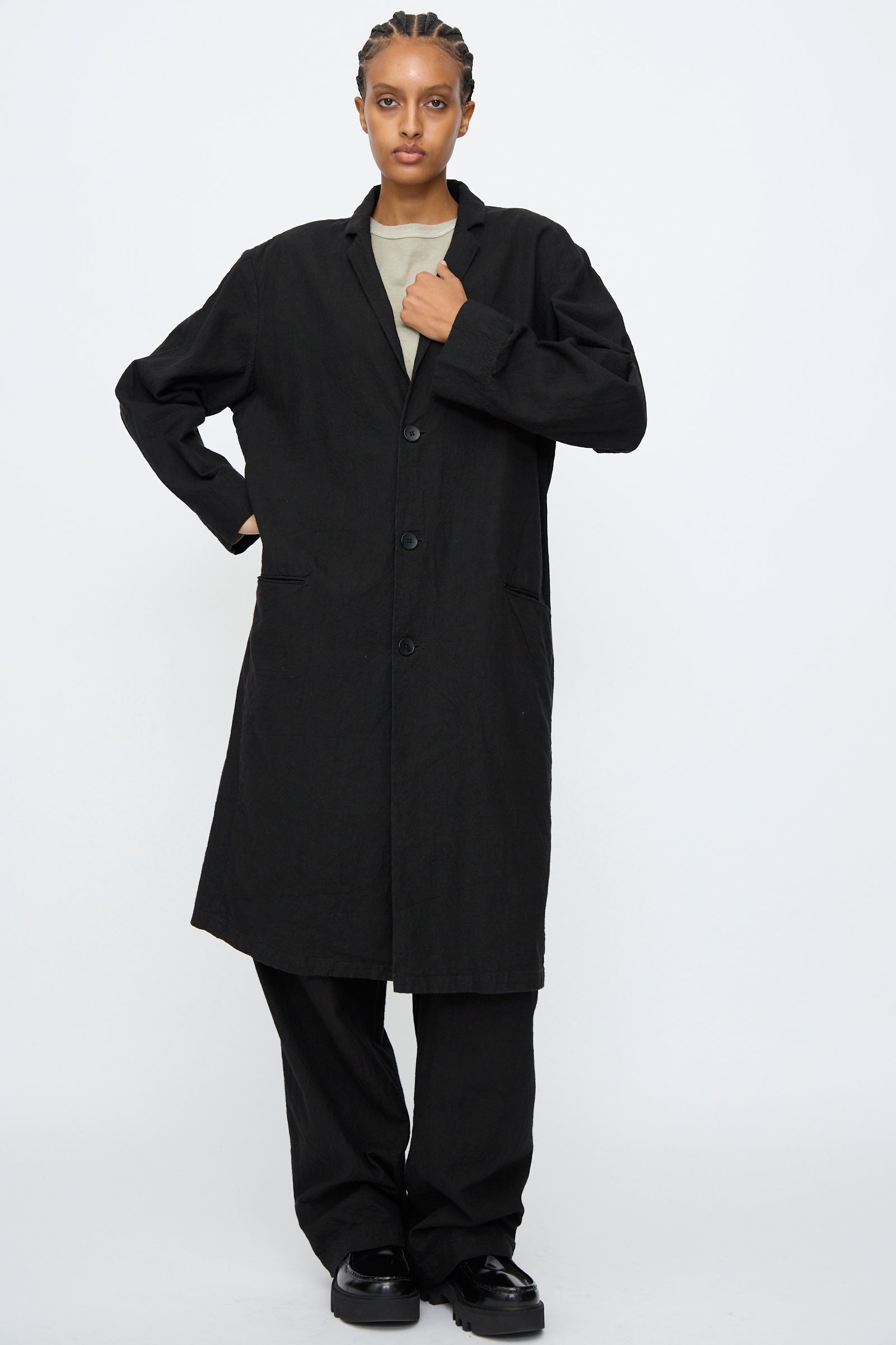 A person wears the Black Crane Crinkled Coat in Black, featuring a subtle crinkled texture, paired with black pants and shoes, standing against a plain white background.